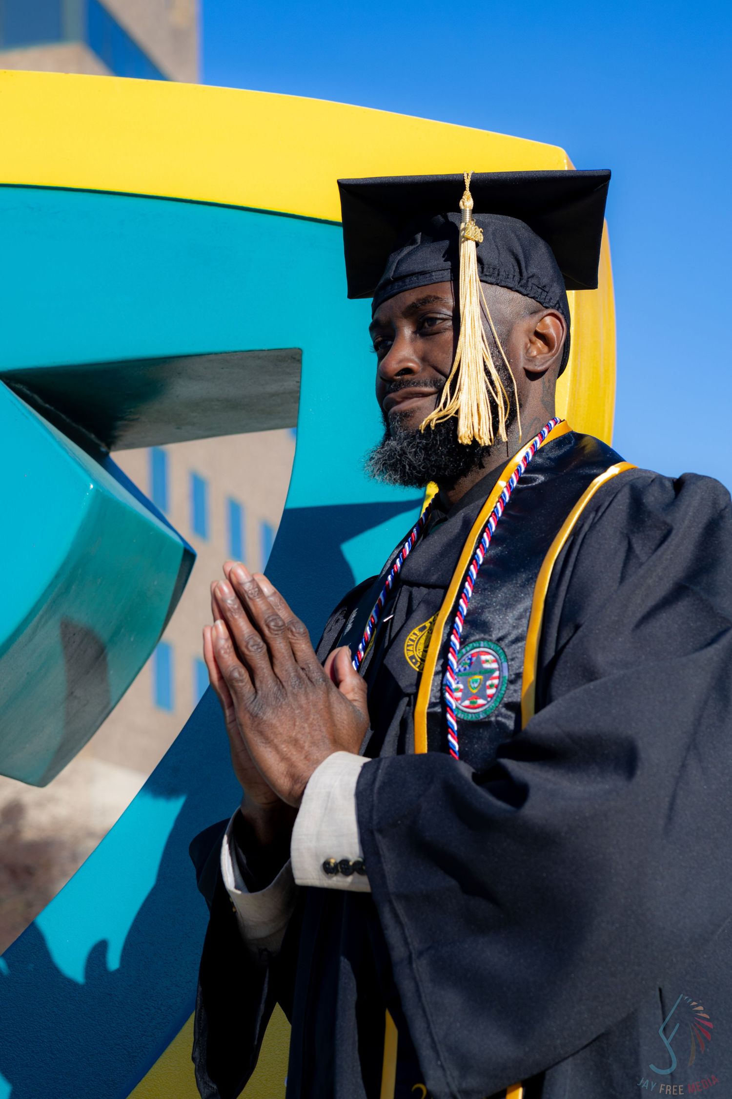 Graduate in cap and gown with honor cords poses proudly at university landmark on a sunny day.