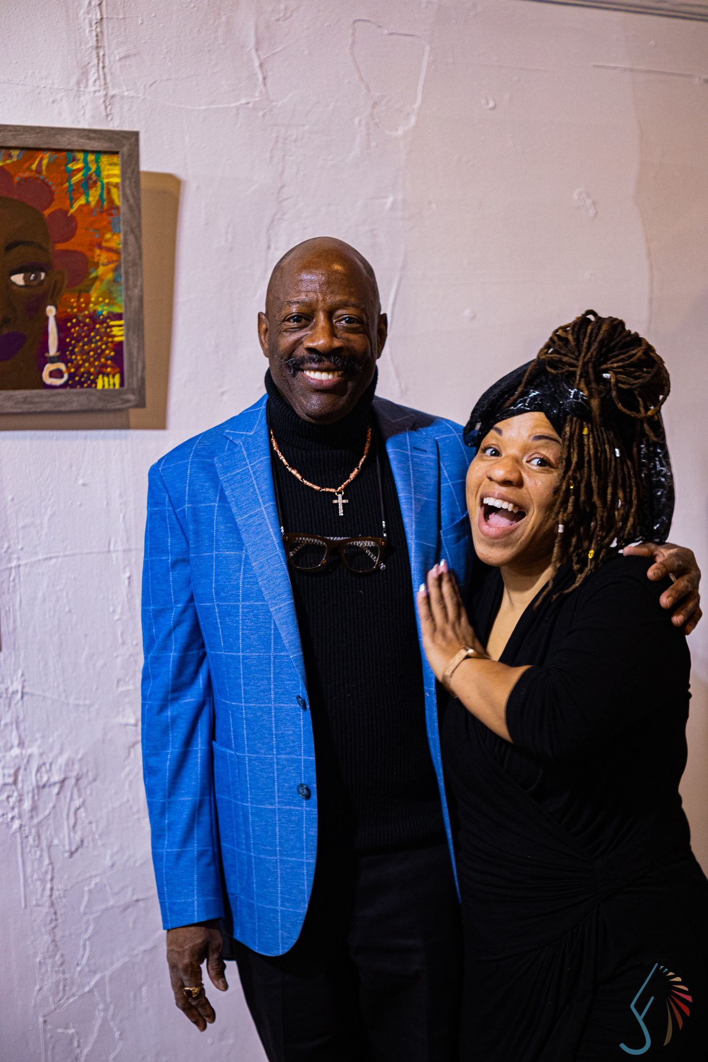 Two smiling people pose together, man in blue blazer with cross necklace and woman in black outfit with dreadlocks, at an art gallery.