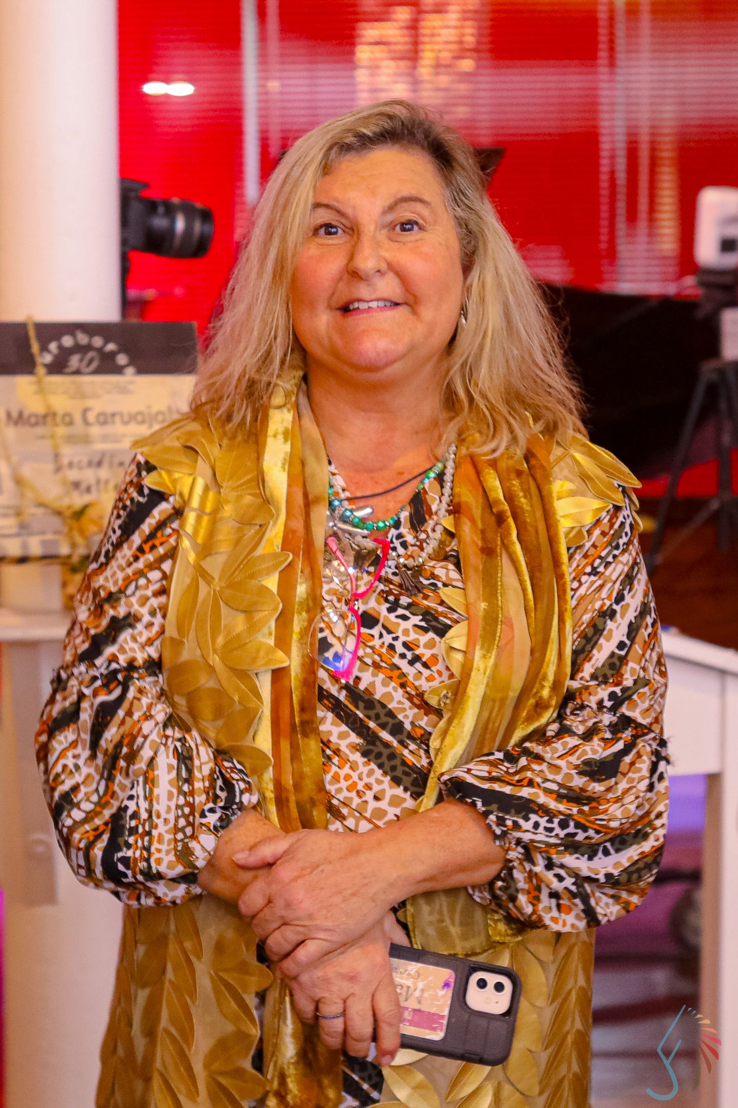 Smiling woman with long blonde hair wearing a gold and animal print blouse, holding a phone at an indoor event.