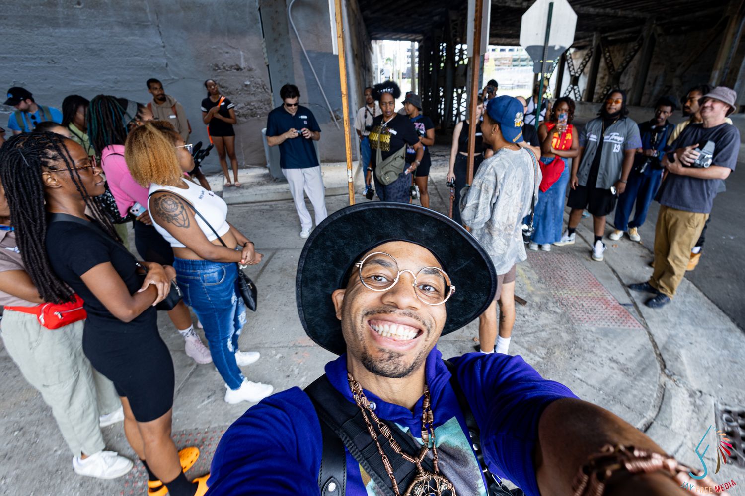 Man takes a selfie smiling under a bridge with a crowd of diverse people gathered behind him.