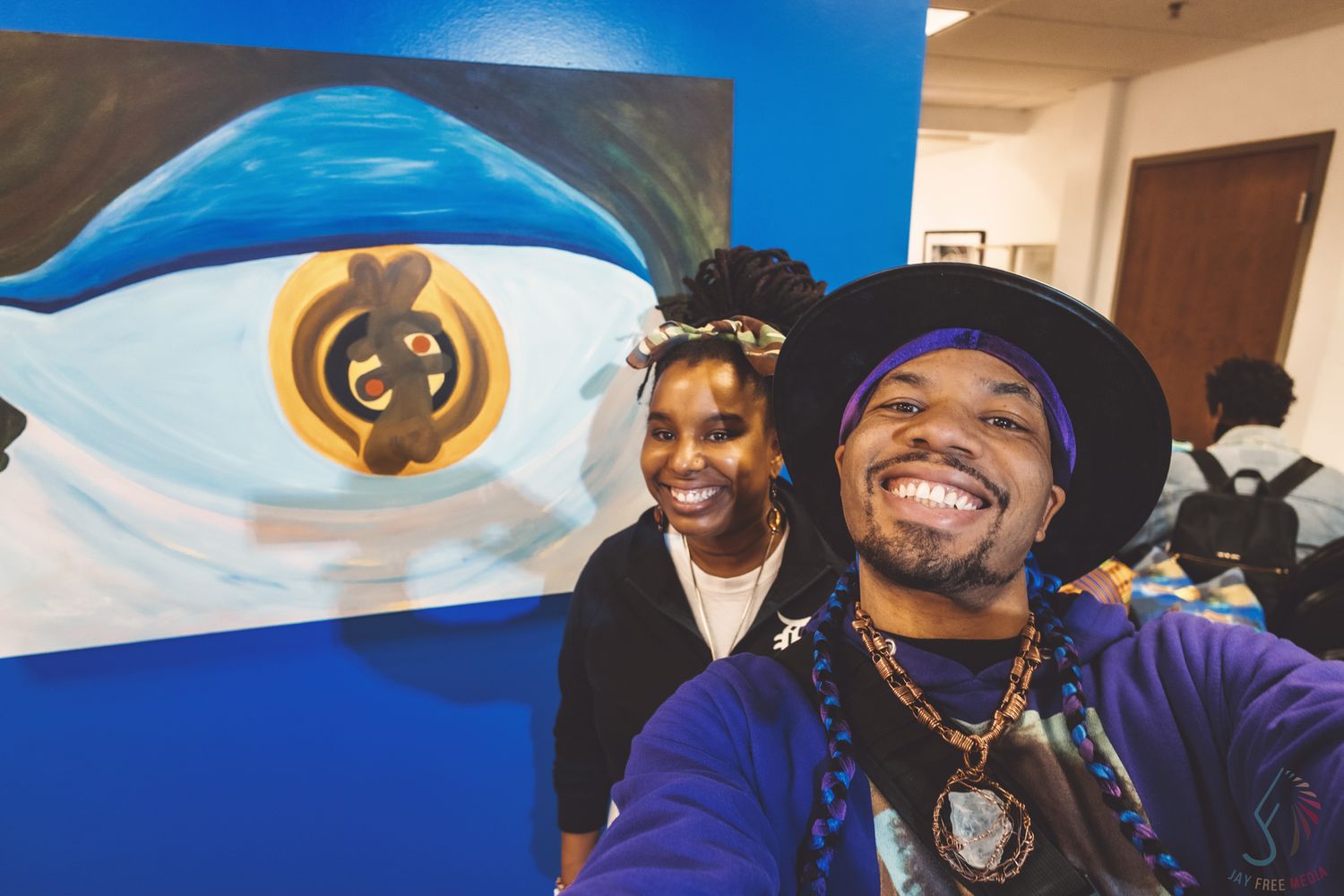 Two people smiling for a selfie in front of a large blue eye painting at an art gallery event.