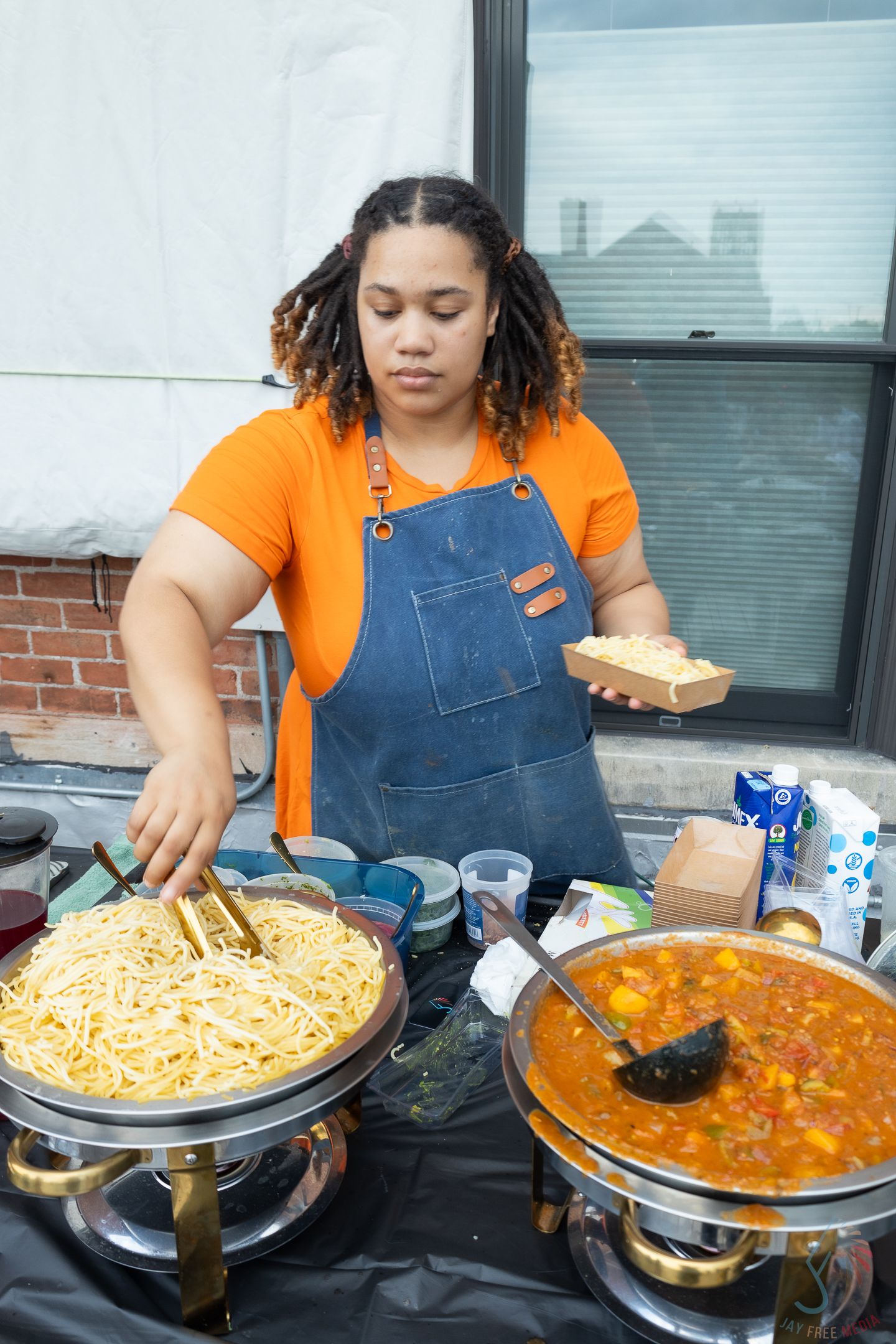 Woman in orange shirt and denim apron serving noodles and curry at an outdoor food stall.