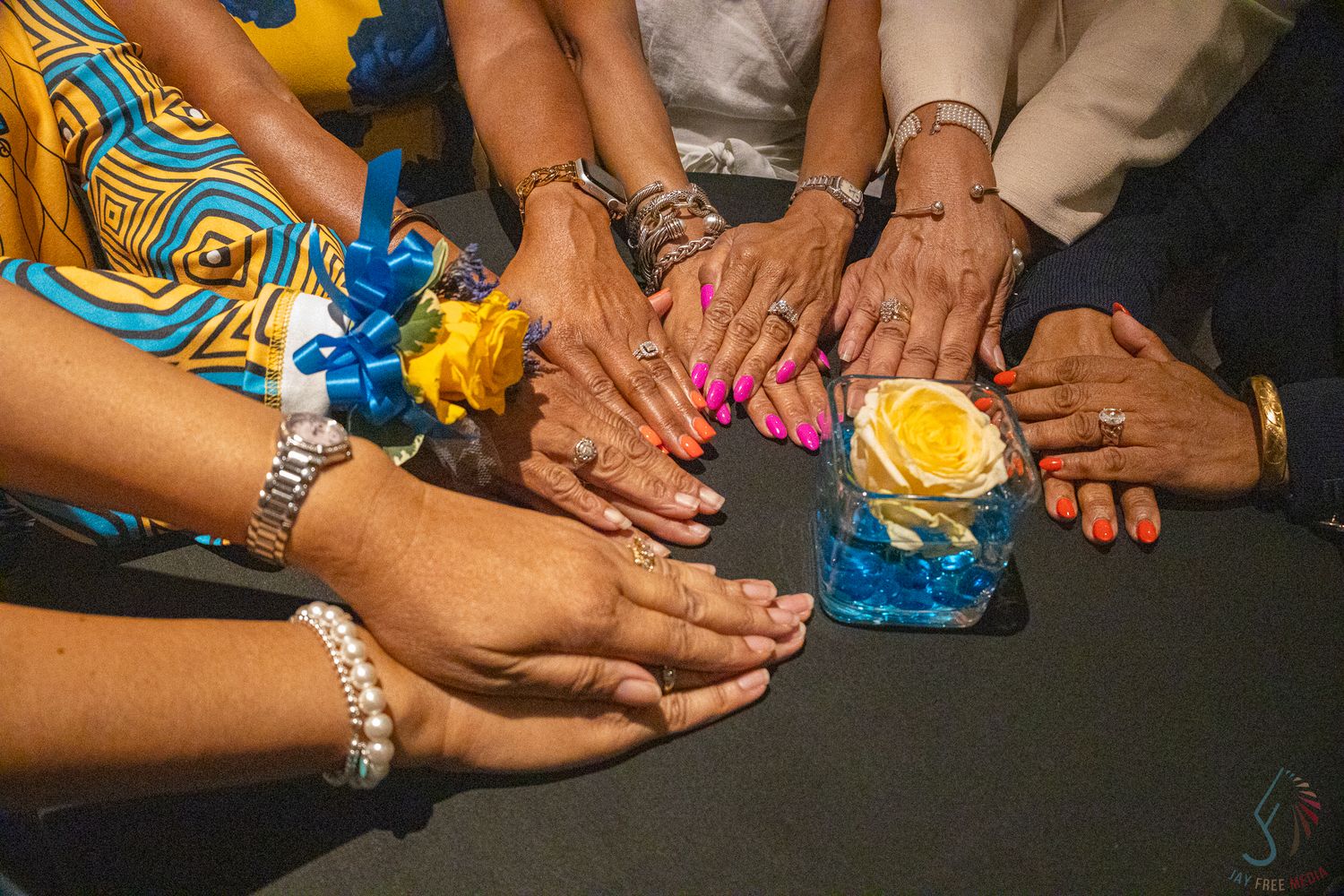 Multiple hands with colorful nail polish and bracelets gathered around a blue vase with yellow rose centerpiece.