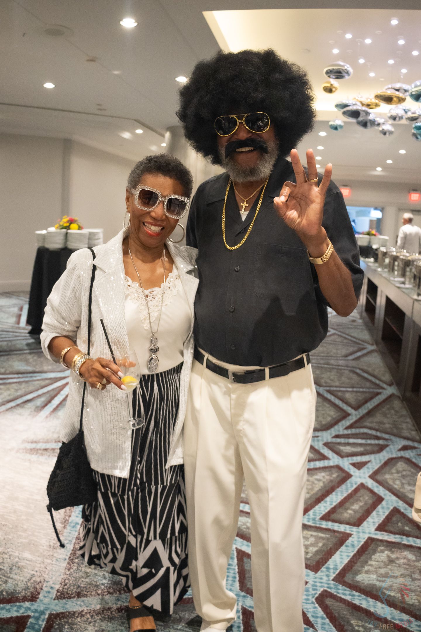 Two people in retro 70s outfits pose at an indoor event, one wearing an afro wig and gold chains.