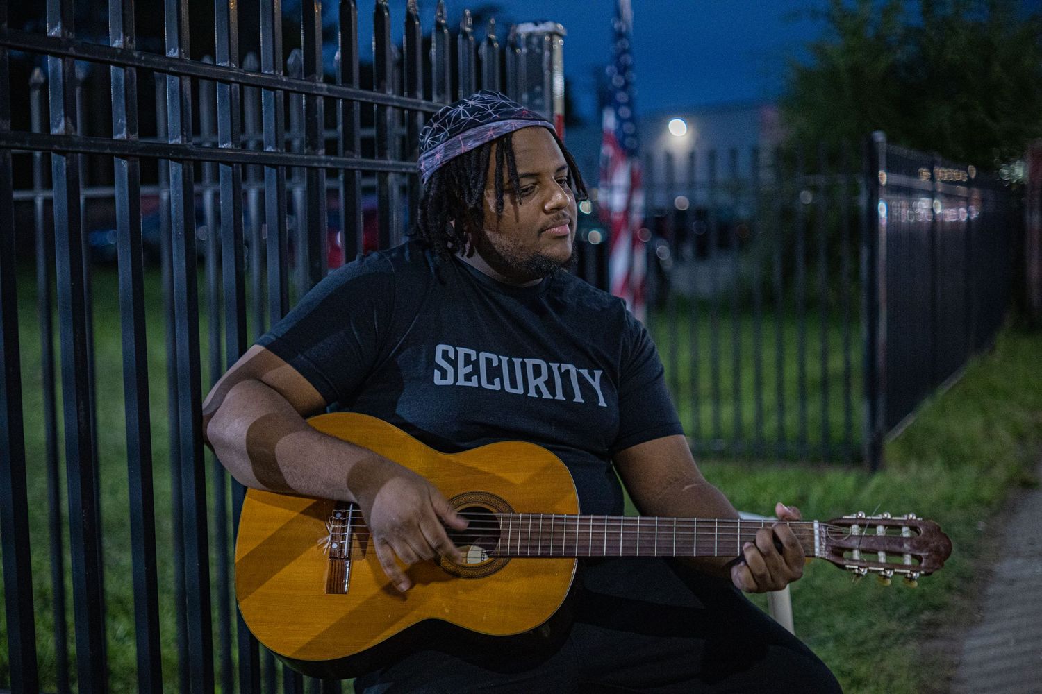 Person in security shirt plays acoustic guitar outdoors near iron fence at dusk.