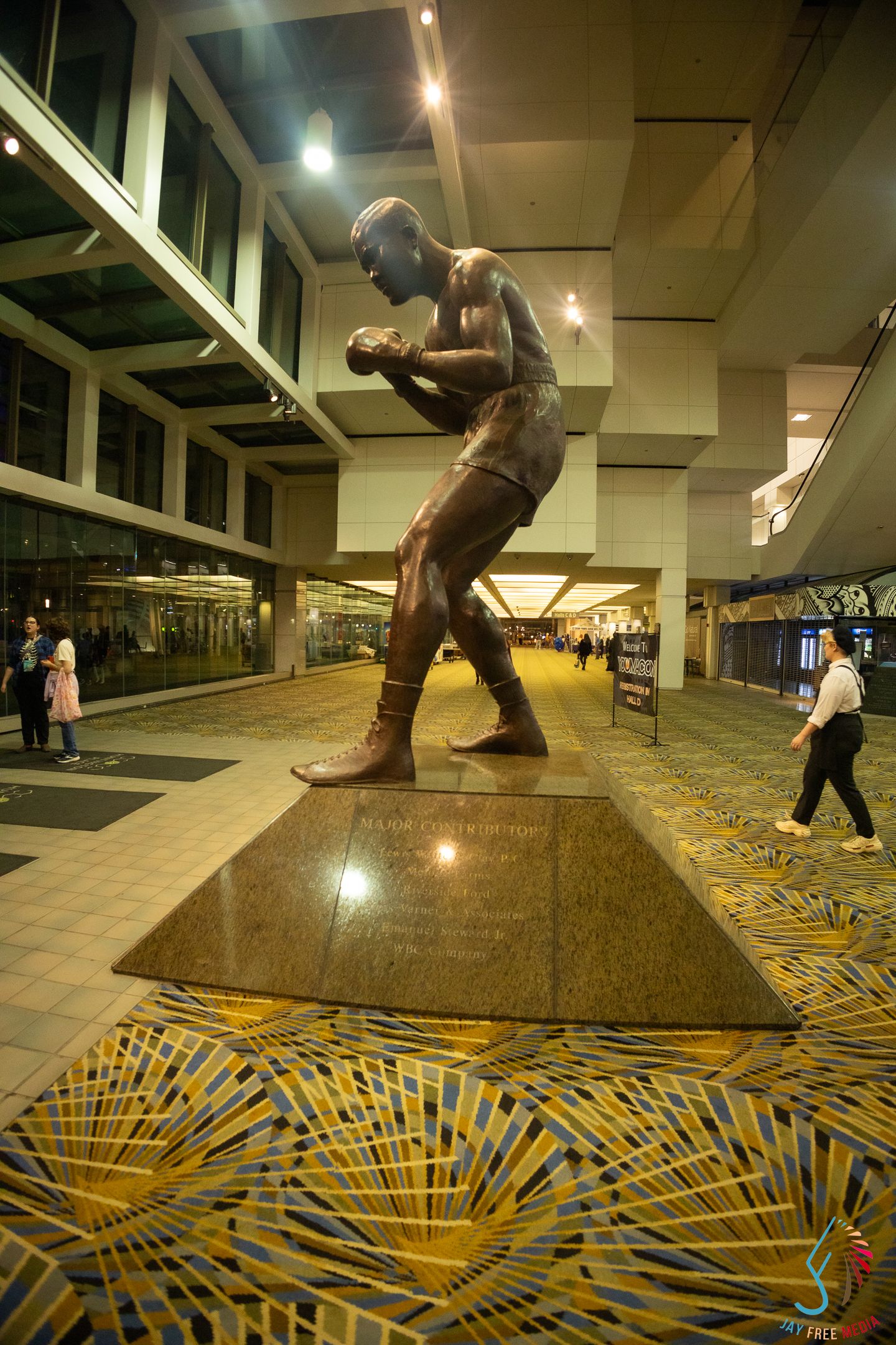 Bronze boxer statue on marble base in modern building lobby with patterned carpet and people walking in background.