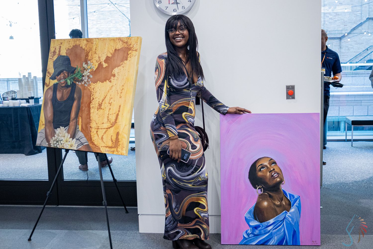 Woman in floral dress poses beside two vibrant portrait paintings at an art exhibition.