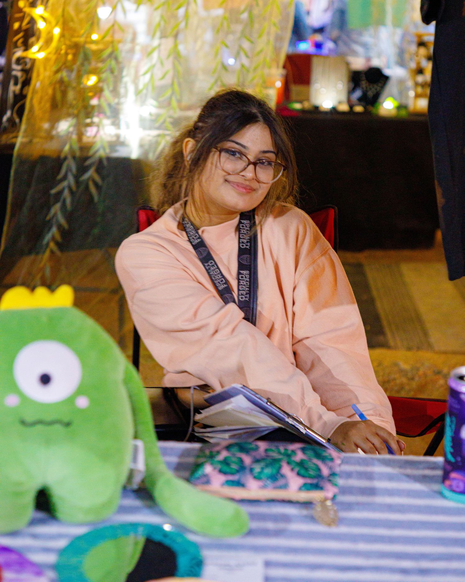 Young woman with glasses smiles at table with green monster plush toy at colorful indoor market event.
