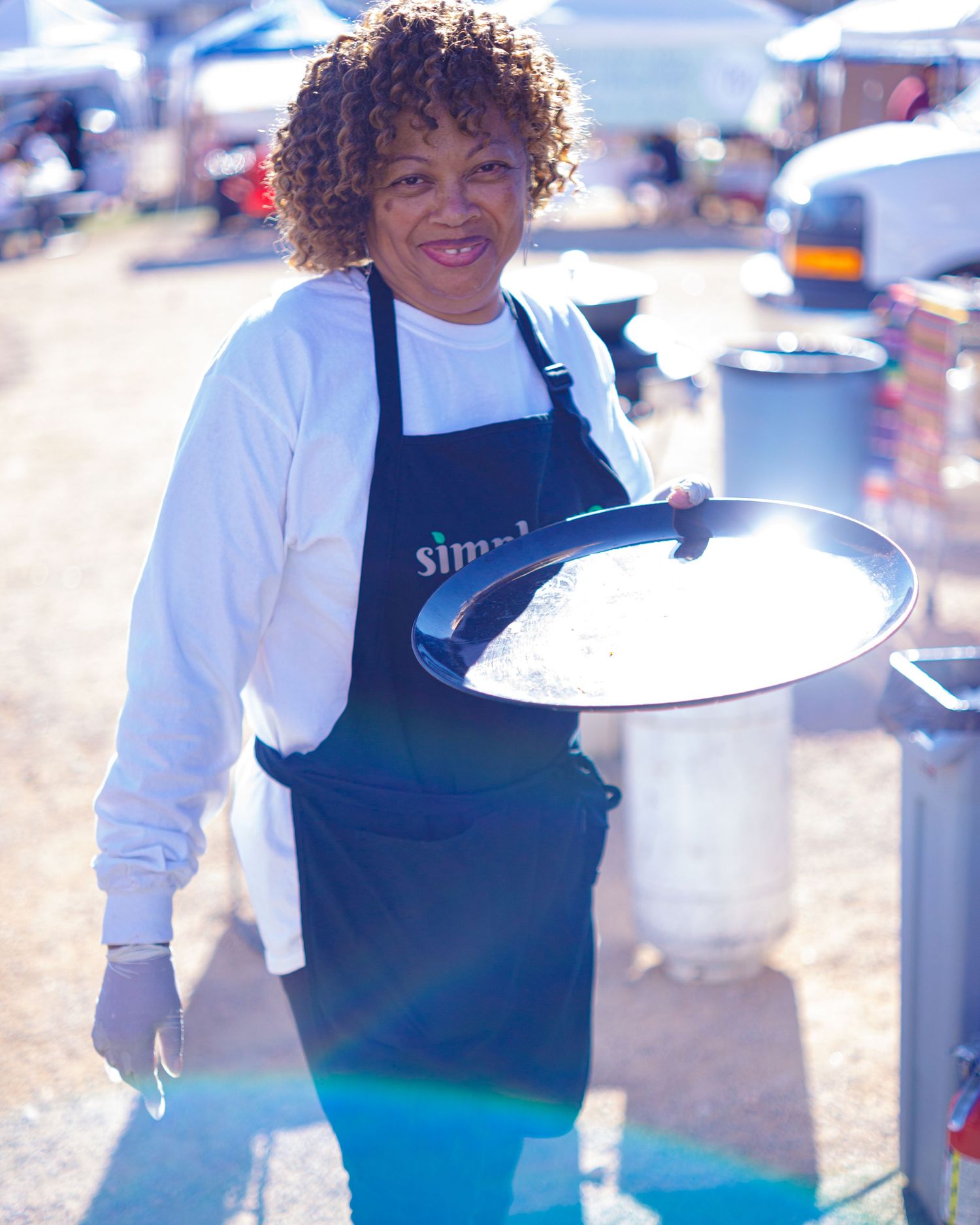 Smiling woman in navy apron holding large pan at outdoor food festival in sunny weather.