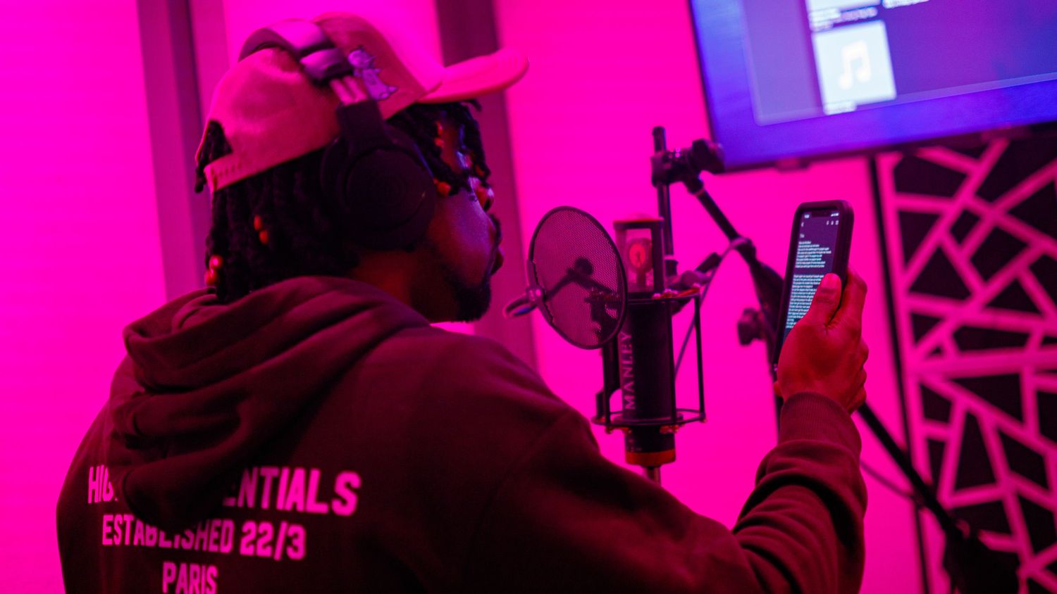 Artist recording in a pink-lit studio, holding a phone near a professional microphone while wearing a hoodie and cap.