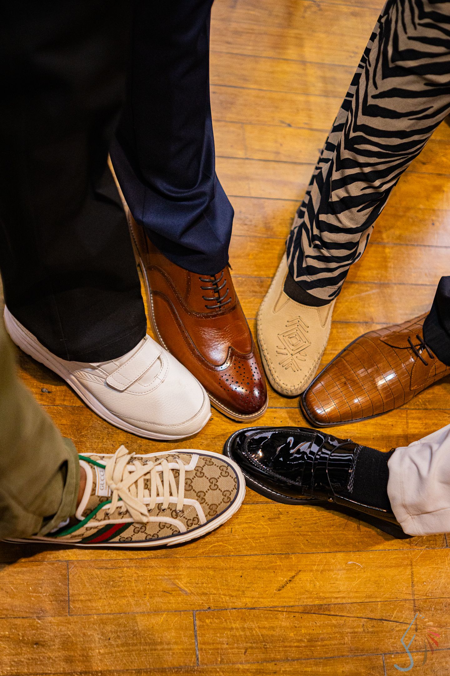 Four pairs of stylish shoes on wood floor, including Gucci sneakers, brown brogues, cowboy boots, and black patent loafers.