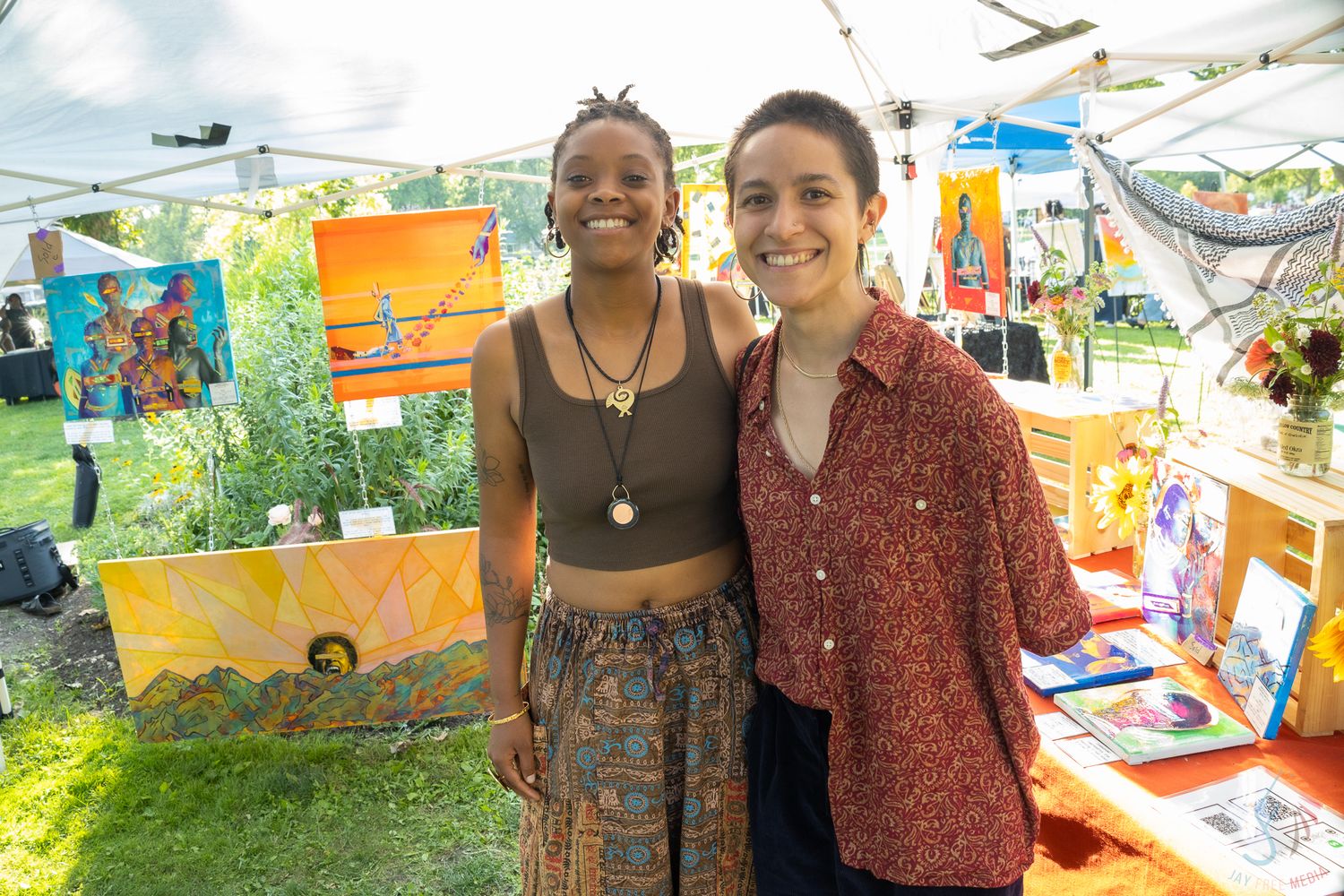 Two young women smiling together at a colorful outdoor art fair booth on a sunny day.