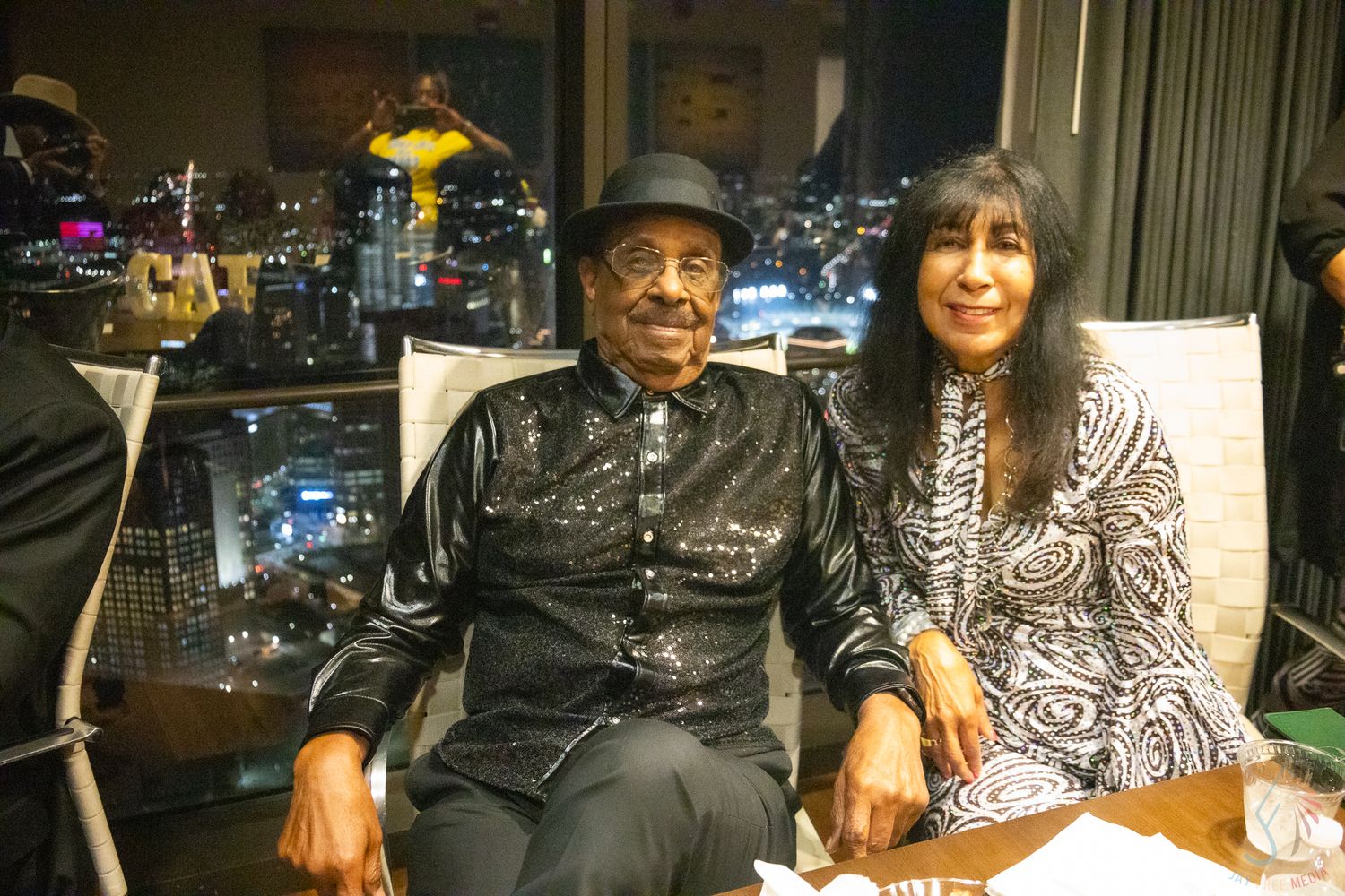 Two people smile together seated indoors with a glowing city skyline visible through the window at night.