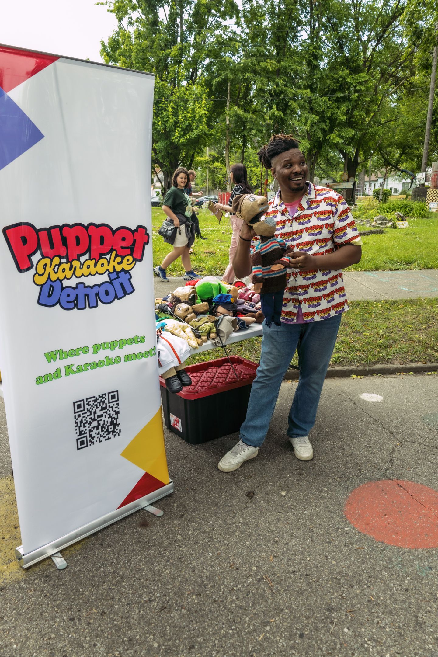 Man standing next to a Puppet Karaoke Detroit banner with puppets at an outdoor community event.