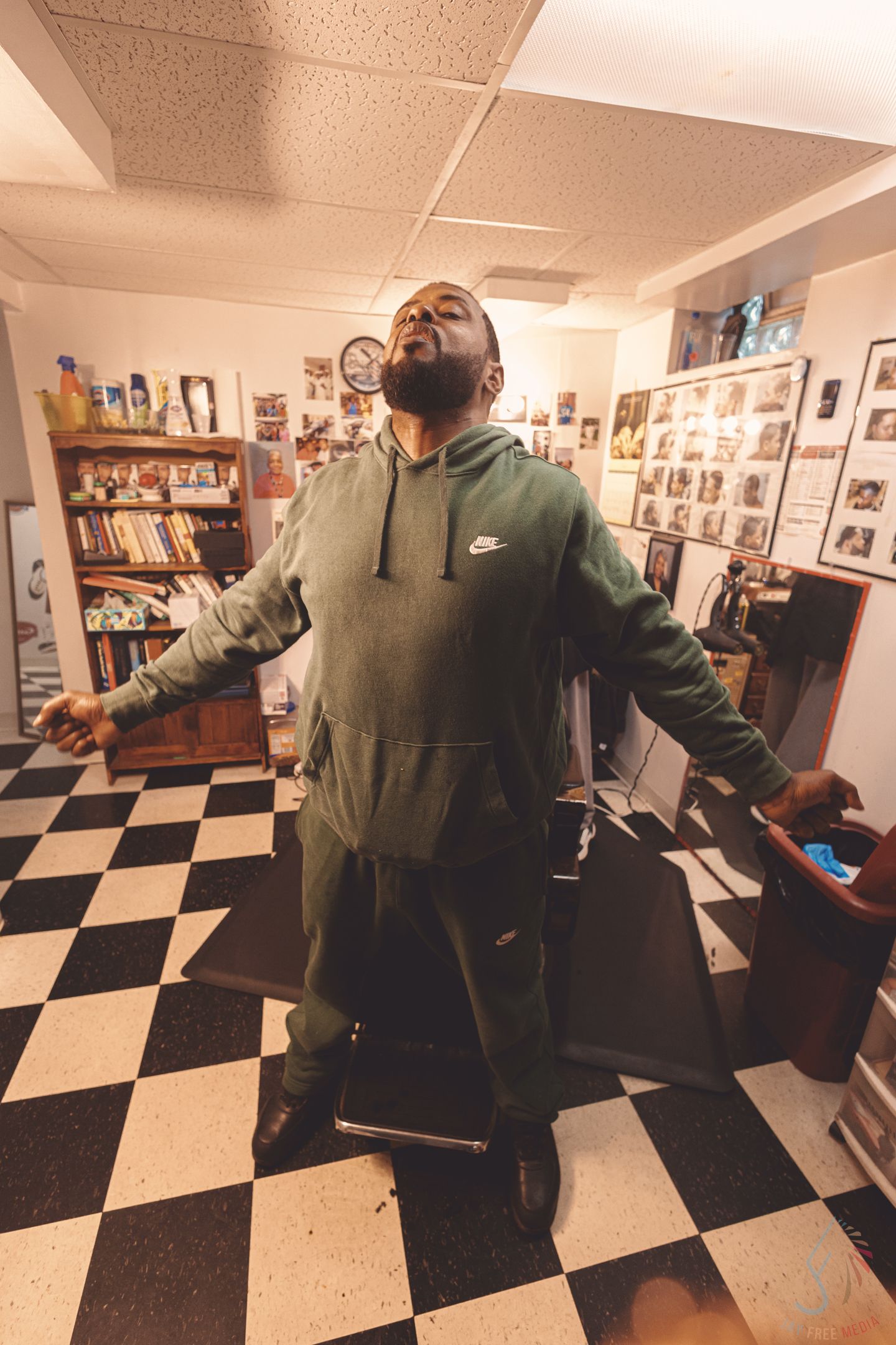 Man in green Nike hoodie and sweatpants poses confidently in a barbershop with checkered floors.