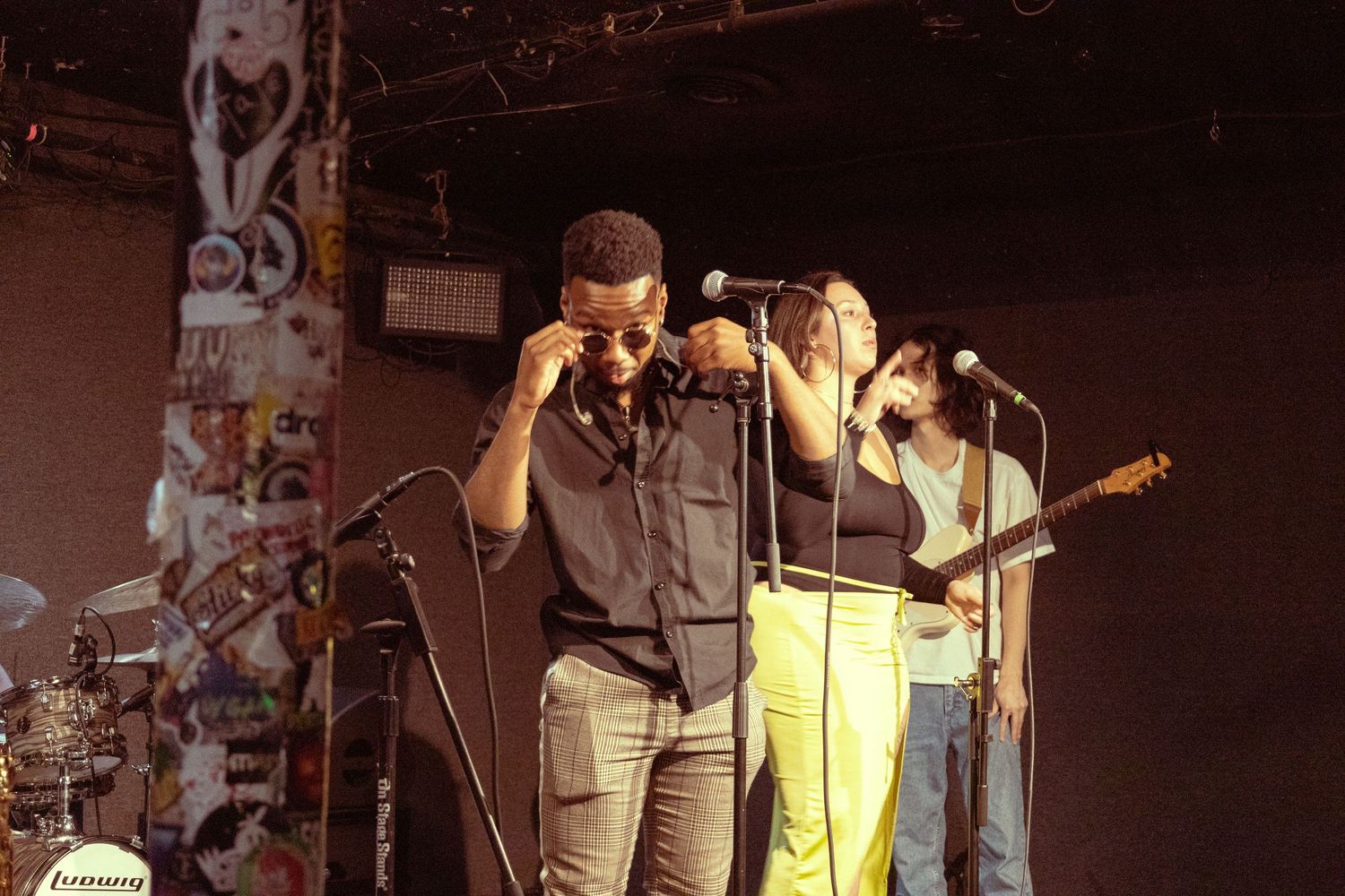 Three musicians performing on stage, with a male singer and female vocalist sharing microphones under warm stage lighting.