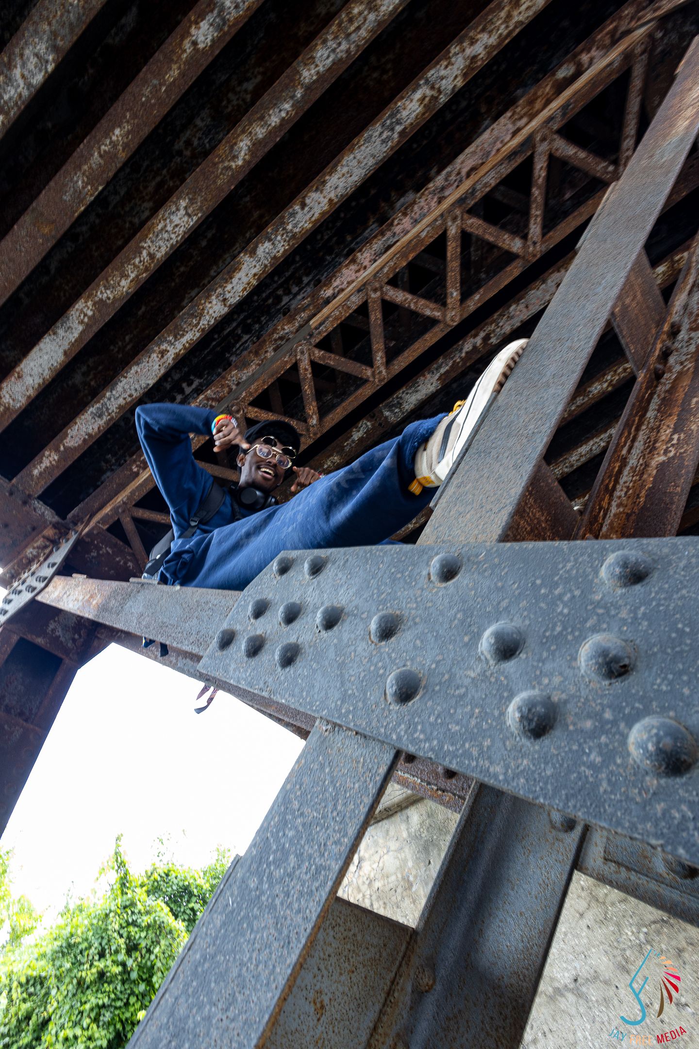 Person resting on steel bridge beams underneath a rusty metal bridge structure outdoors.