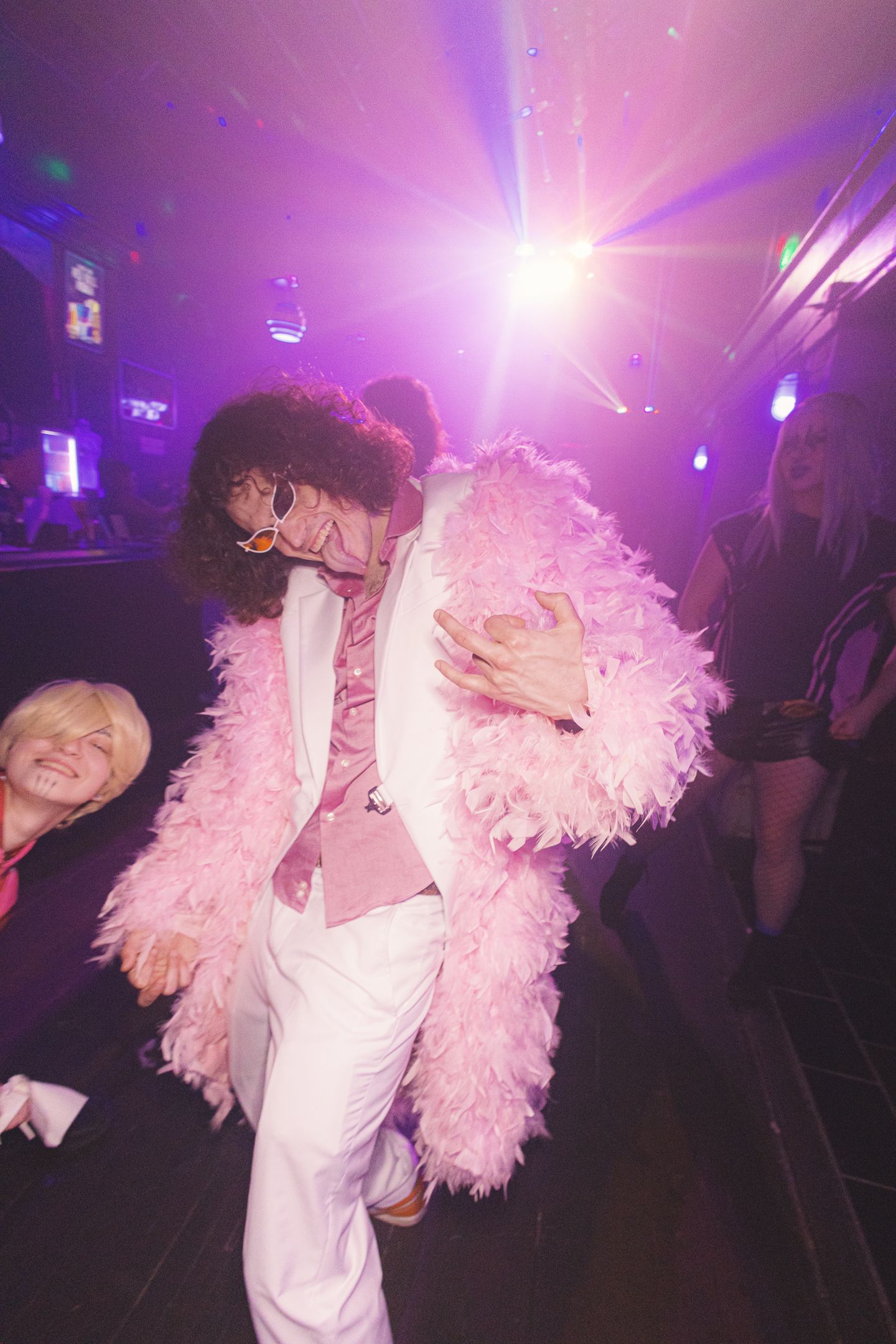 A person in a pink feathered coat and white suit dances under purple disco lights at a nightclub party.