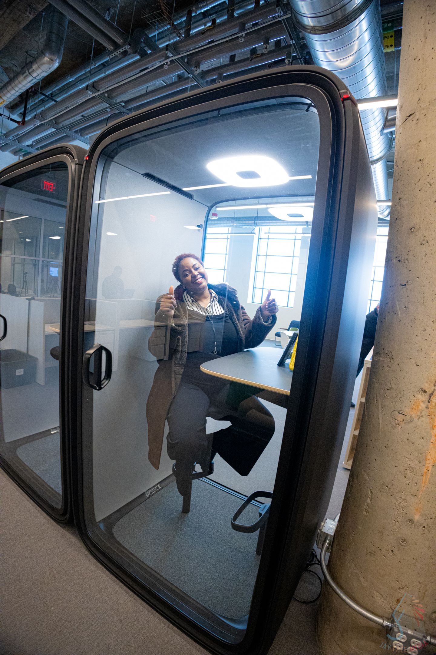 Woman smiling and talking on phone inside a modern office privacy pod with glass door and built-in desk.