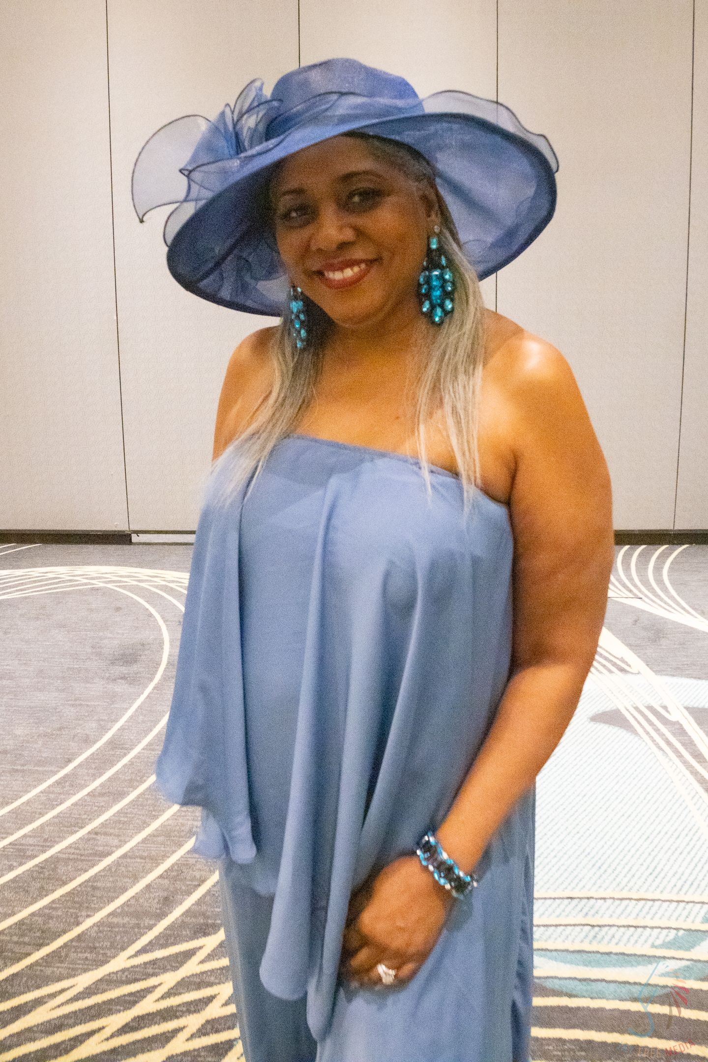 Woman in blue strapless dress and wide-brim hat with teal earrings and bracelet smiling at formal event.