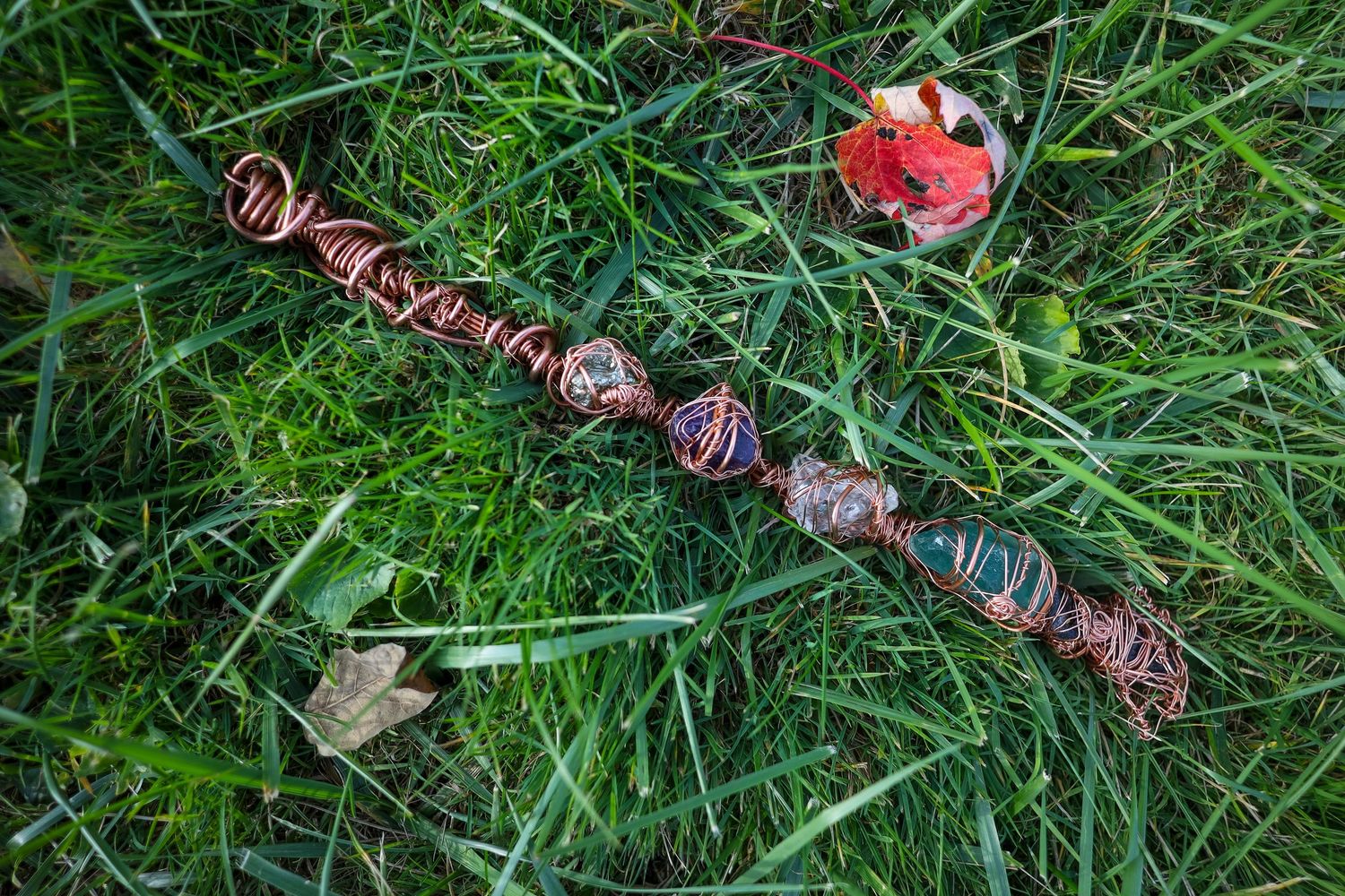 Handcrafted copper wire wrapped wand with colorful beads laying on green grass, perfect for fantasy or Wiccan rituals.