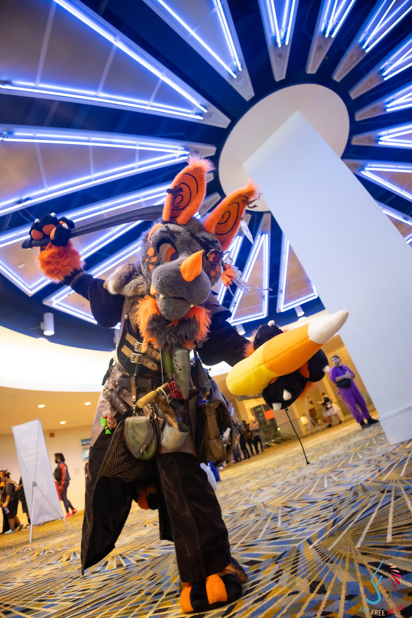 Fursuit cosplayer in elaborate gray and orange animal costume poses in convention center lobby with neon light display.