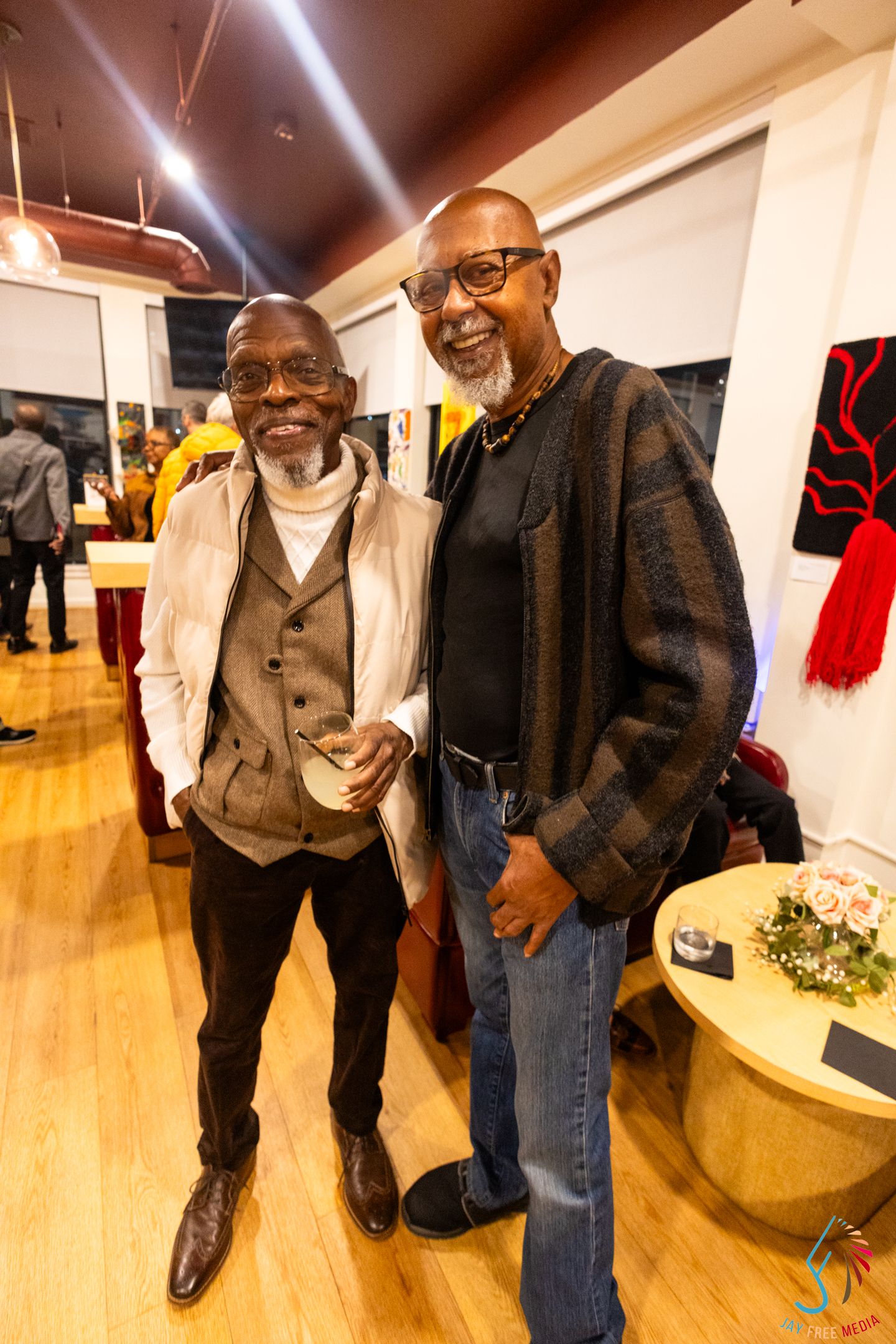 Two smiling men pose together at an art gallery social event on hardwood floors.