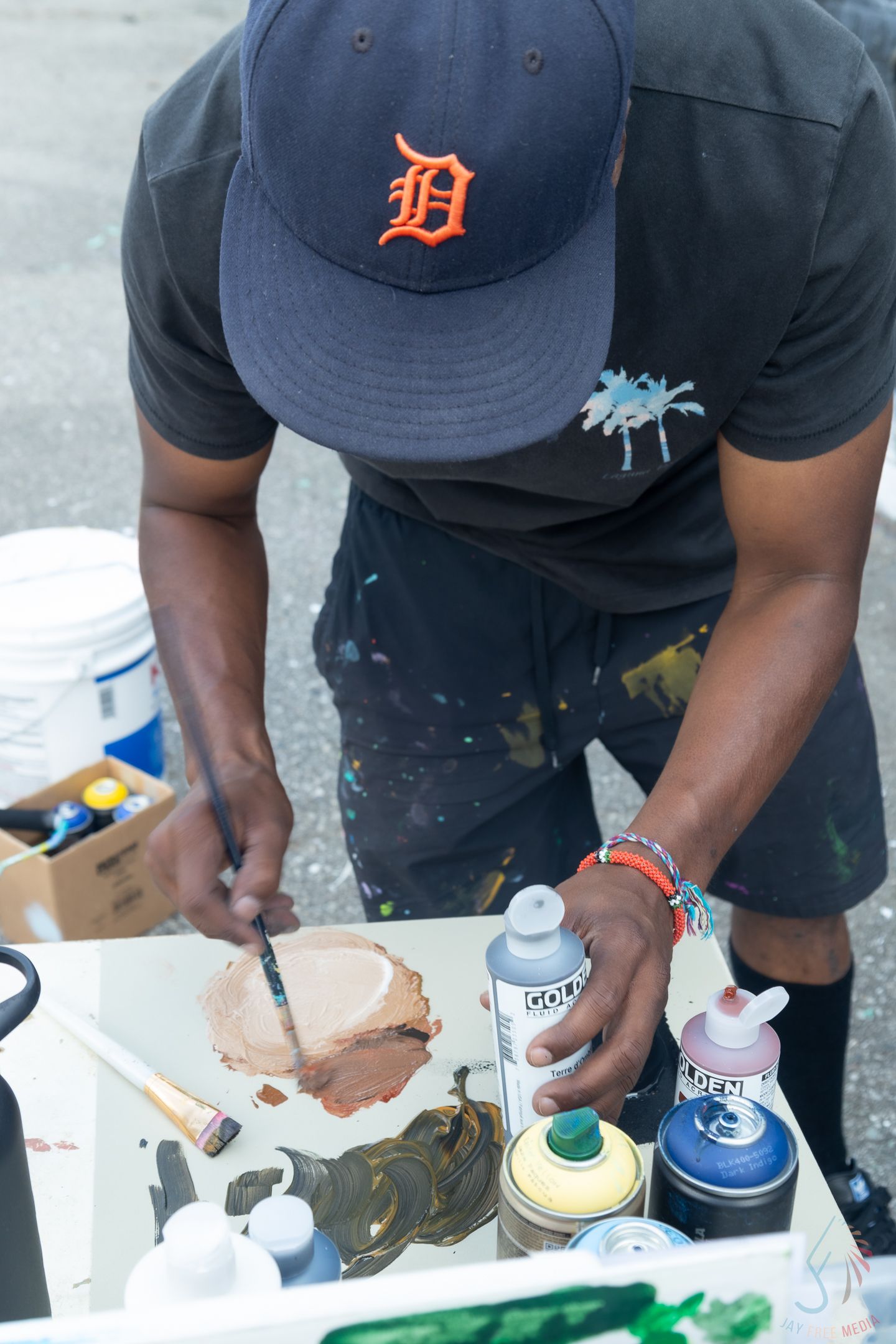 Artist in Detroit Tigers cap mixing brown paint with brush and spray cans on outdoor table.