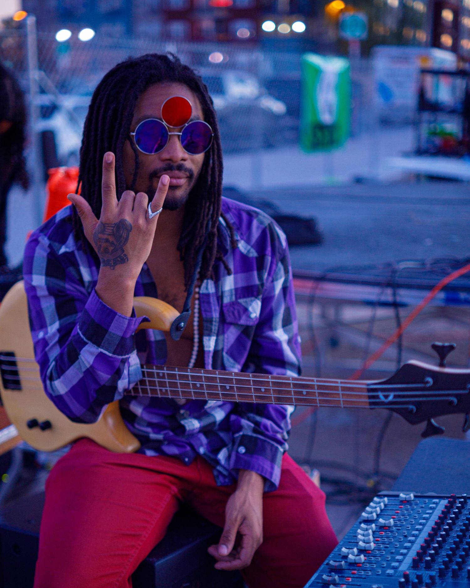 Musician with dreadlocks and round tinted glasses plays bass guitar at outdoor evening event.