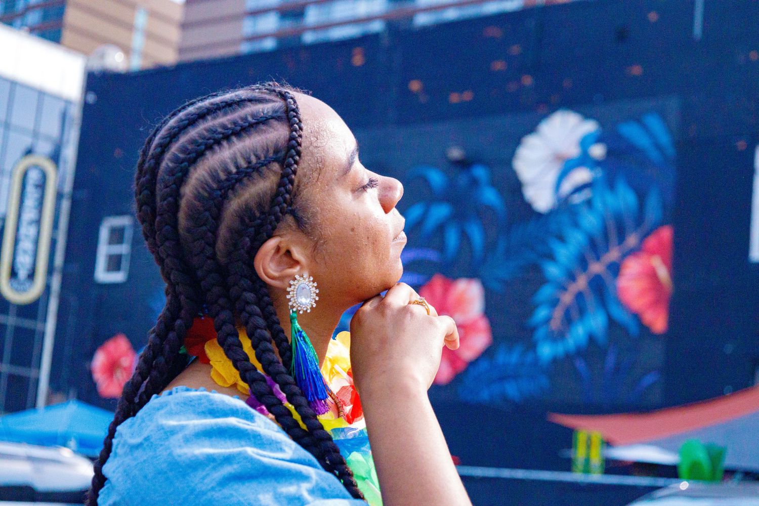 Young Black woman with colorful braids gazes upward thoughtfully in front of a vibrant tropical floral mural.