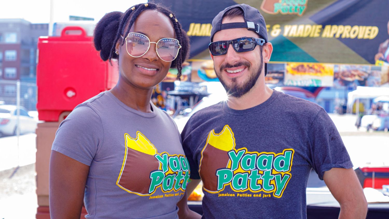 Two smiling people wearing Yaad Patty branded t-shirts at an outdoor Jamaican food event on a sunny day.