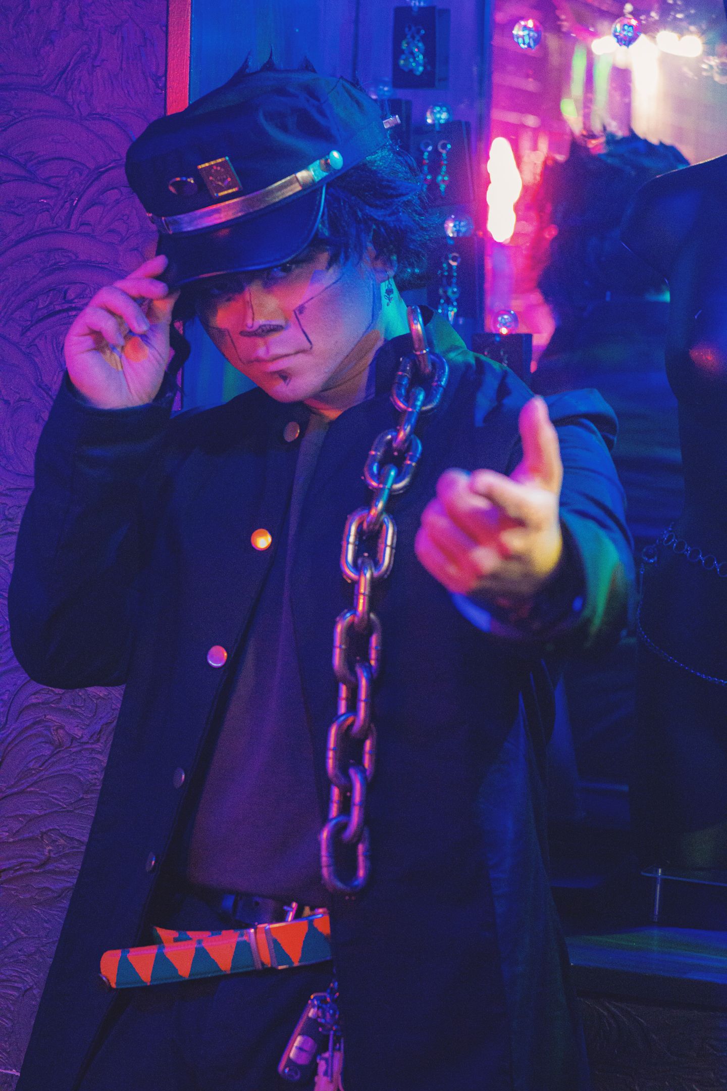 Person in dark uniform with cap and large chain accessory posing under neon lights in a dramatic cosplay outfit.