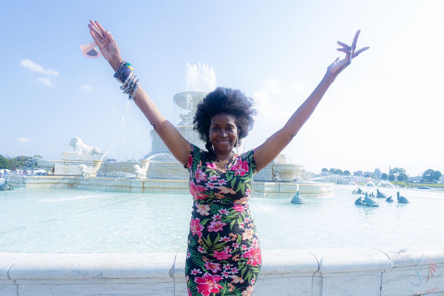 Joyful woman in floral dress with arms raised in front of a large ornate fountain on a sunny day.