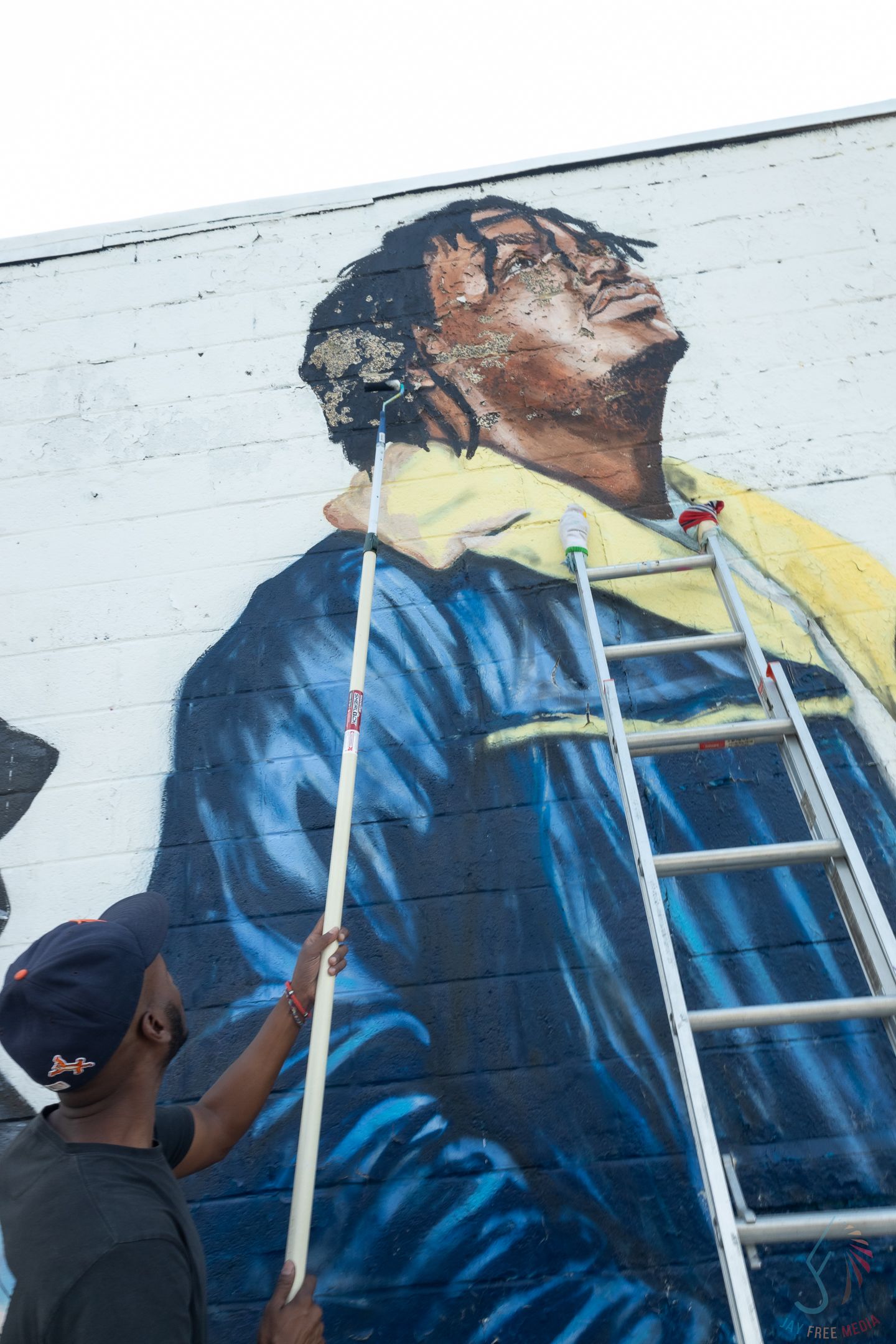 Artist painting a large mural of a man in blue and yellow jacket on a brick wall using a ladder and long brush.