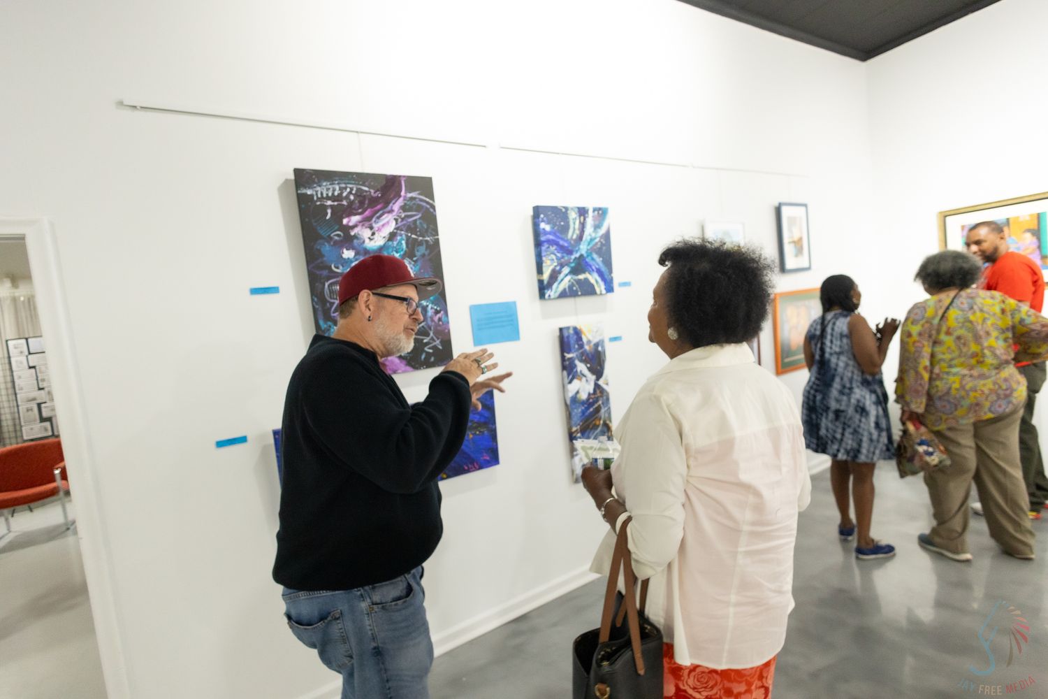 Art gallery visitors discussing colorful paintings on white walls during an exhibition opening event.