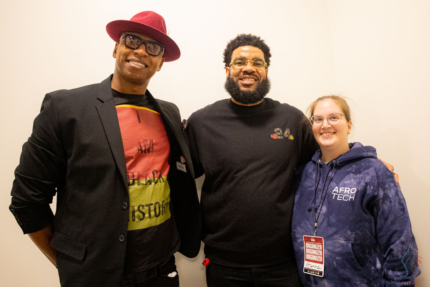 Three people smiling together at Afro Tech conference, one wearing a red hat and colorful shirt, another in a black hoodie.