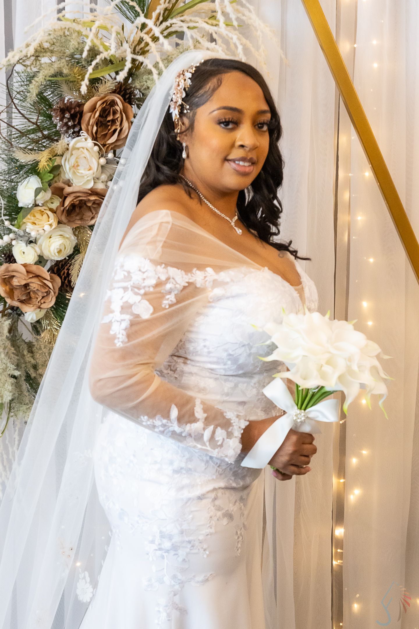 A bride in a white lace mermaid gown with sheer sleeves holds a bouquet, wearing a veil and floral headpiece.