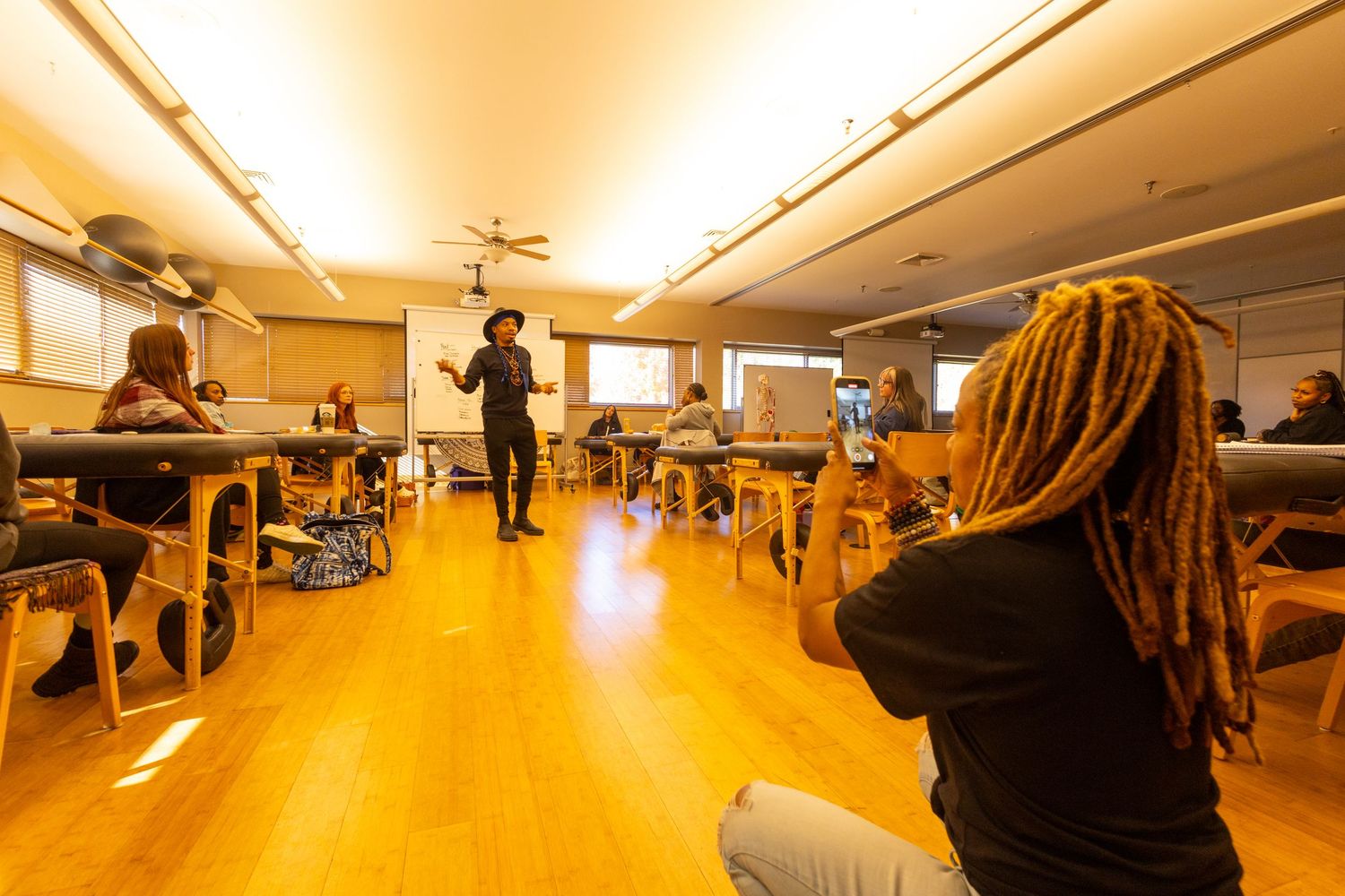 Person in black hat presenting to a group of students seated in a warmly lit classroom with wooden floors.