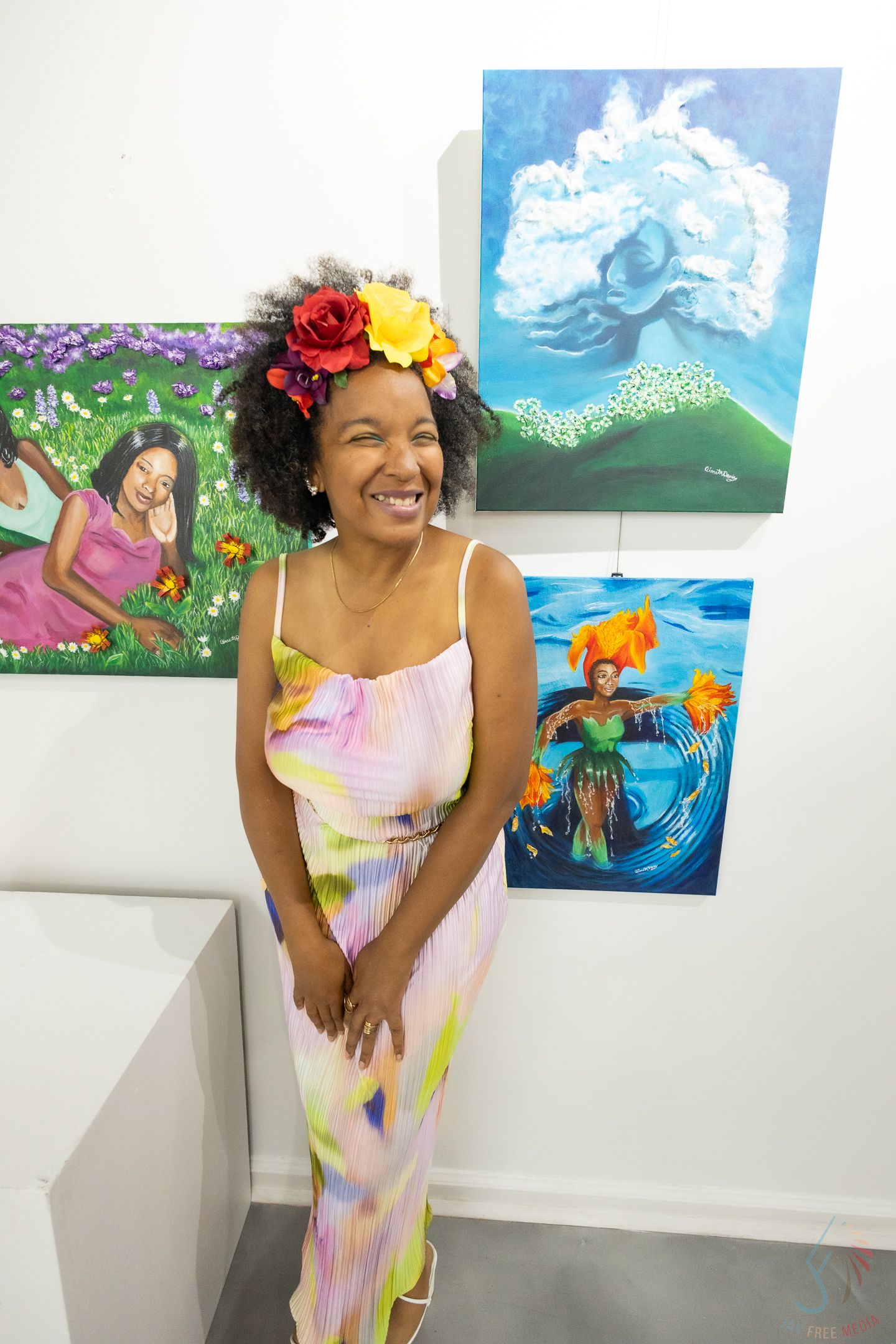 Smiling woman wearing colorful floral crown and pastel dress standing in front of vibrant paintings at an art gallery.