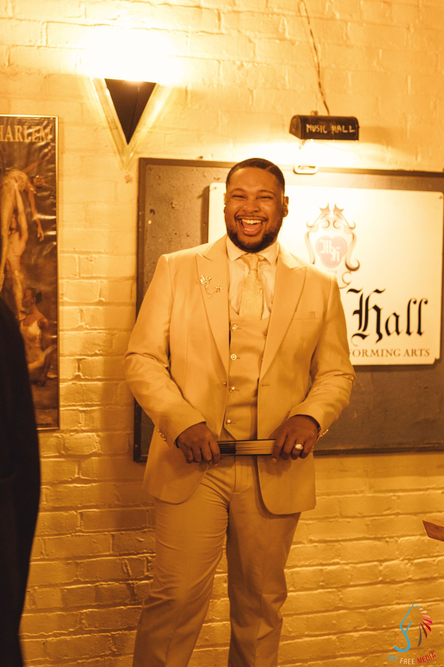 Smiling man in a tan three-piece suit standing in front of a Hall Performing Arts sign on a brick wall.