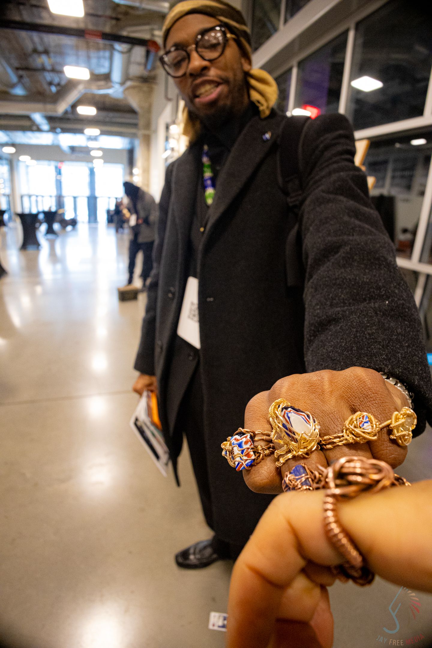Man wearing glasses and black coat displays ornate gold and gemstone rings and bracelets at a jewelry showcase.