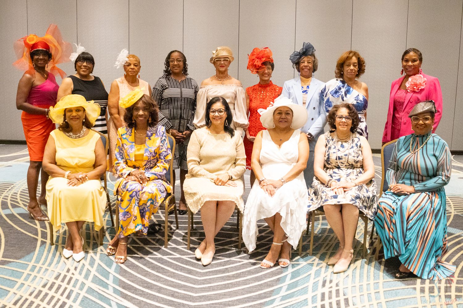 Group of elegantly dressed women wearing colorful hats posing together for a formal group photo at an event.
