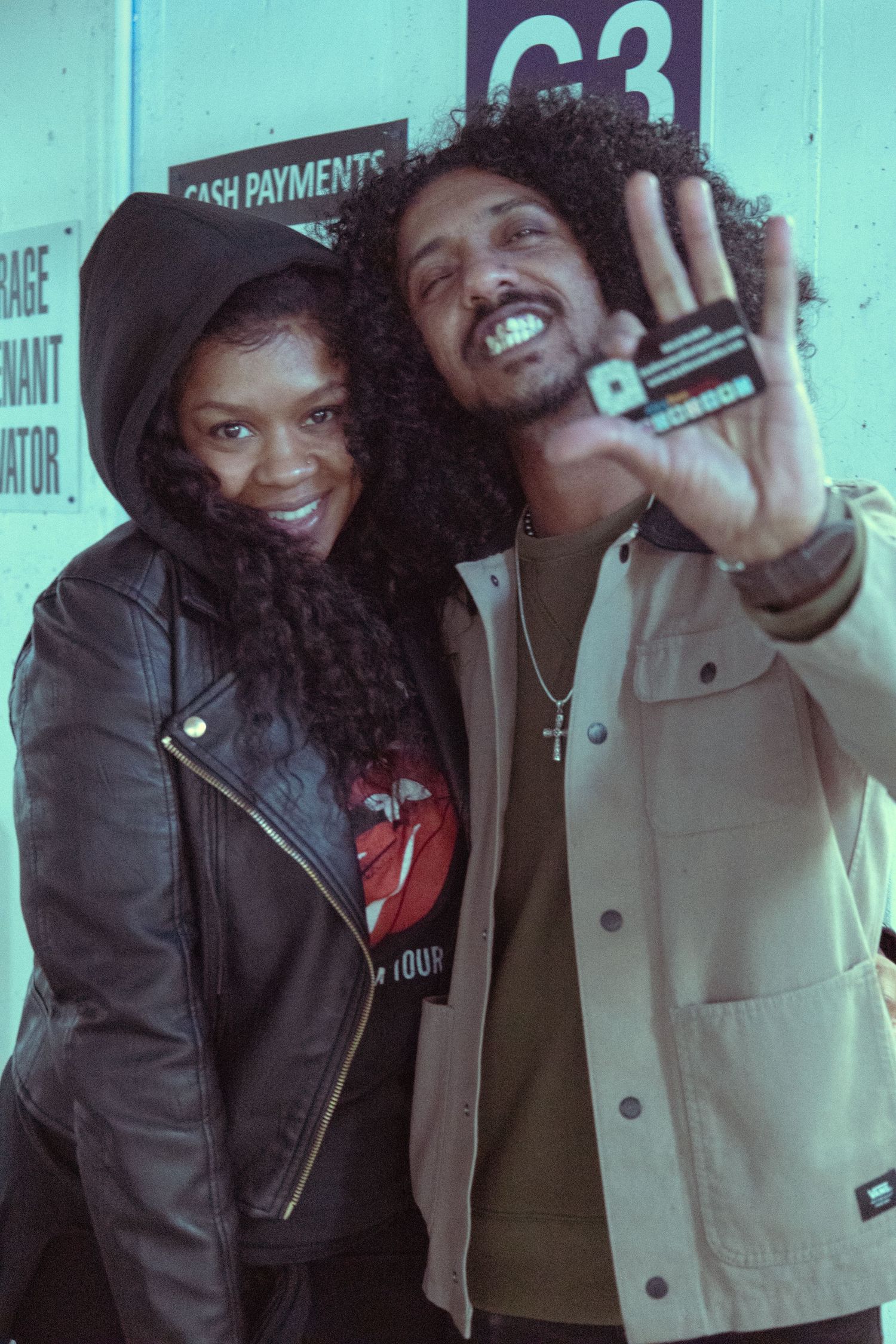 Two friends posing together near a G3 cash payments sign, woman in leather jacket and man in beige jacket holding up a card.