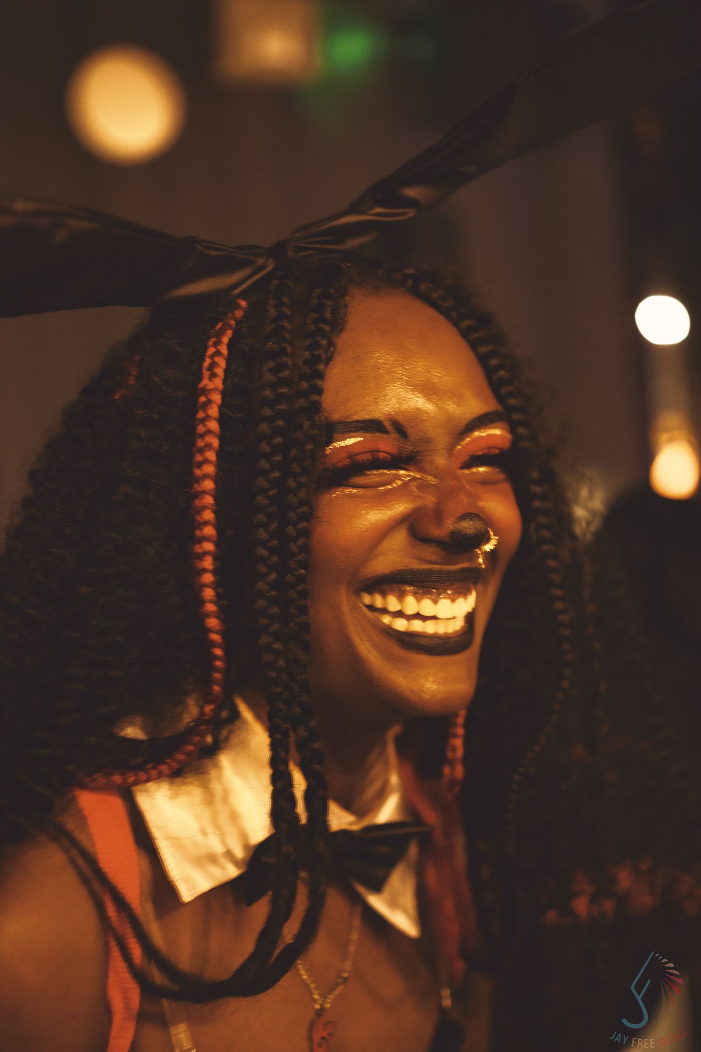 Joyful Black woman with braids, nose ring, and bold eye makeup laughing at a warmly lit event.