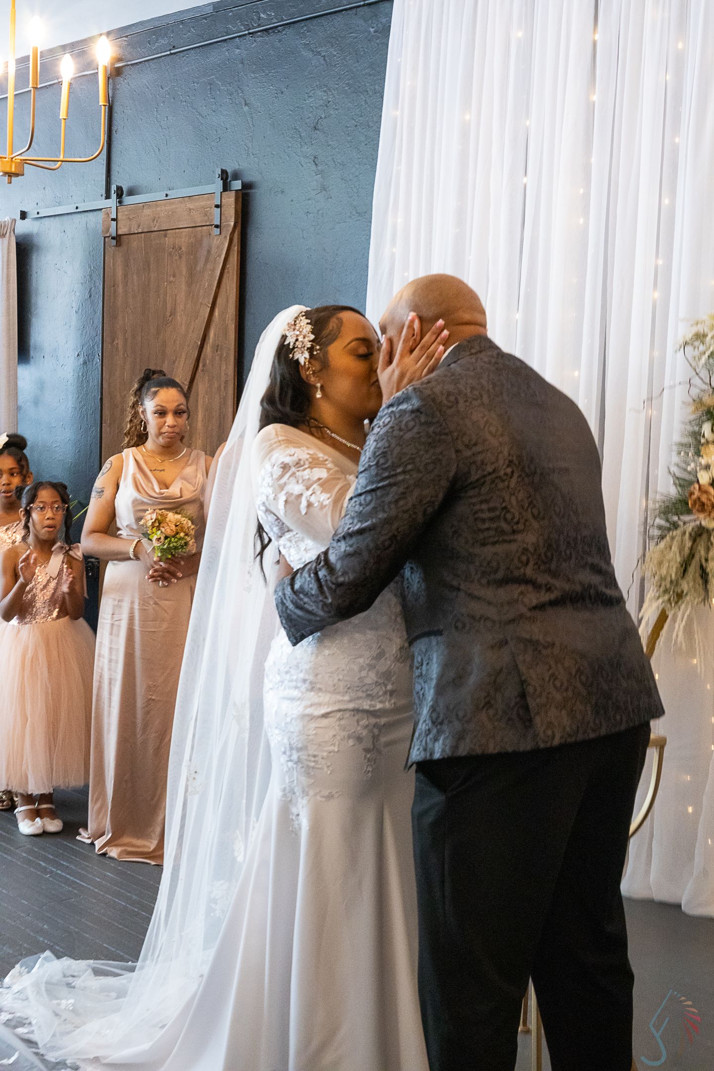 Bride and groom share their first kiss at indoor wedding ceremony with bridesmaids in blush gowns standing nearby.