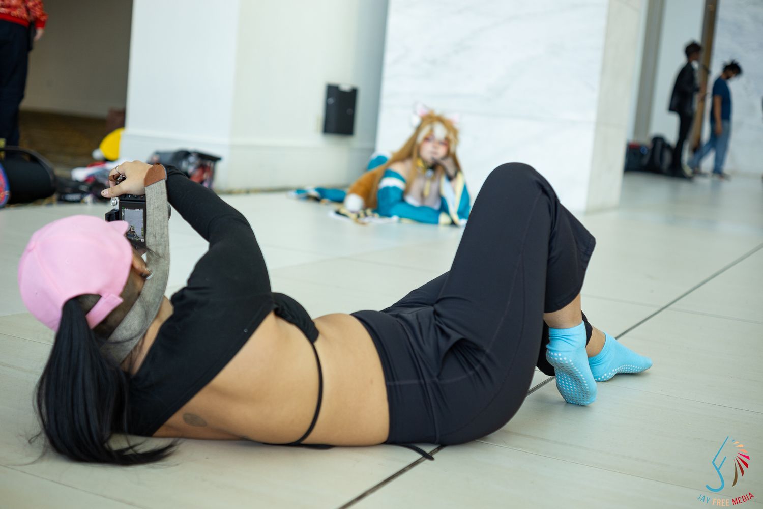 Cosplayer in black outfit blows pink bubble gum while lying on convention floor using a tablet.