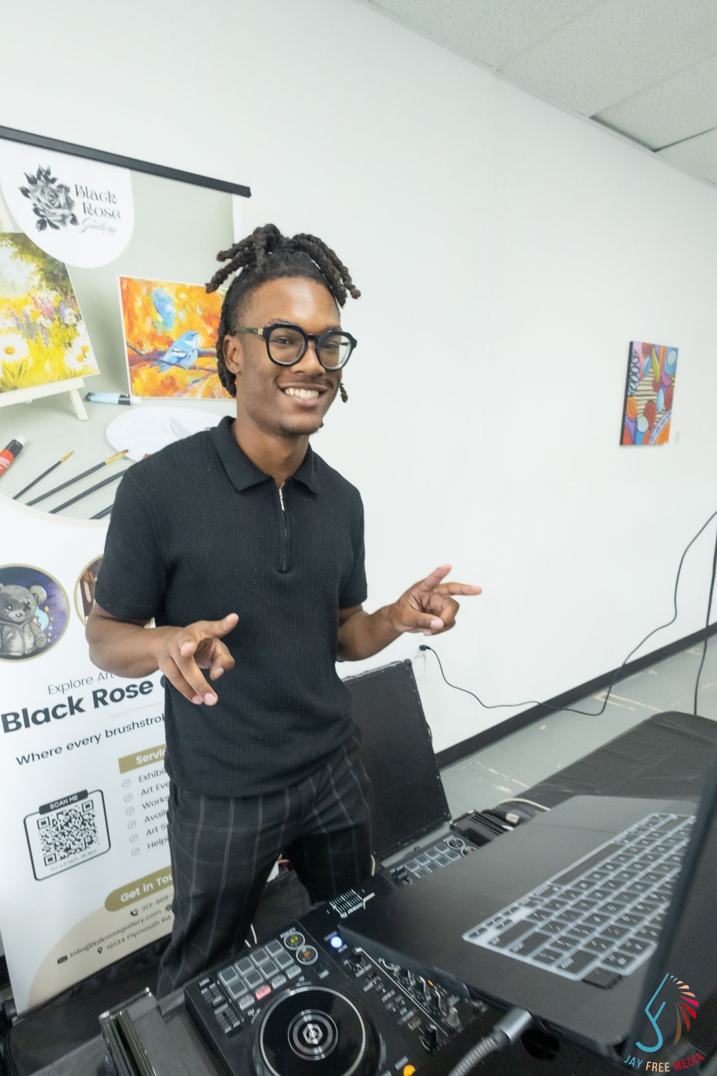 DJ smiling and pointing at camera behind turntables at Black Rose art event.