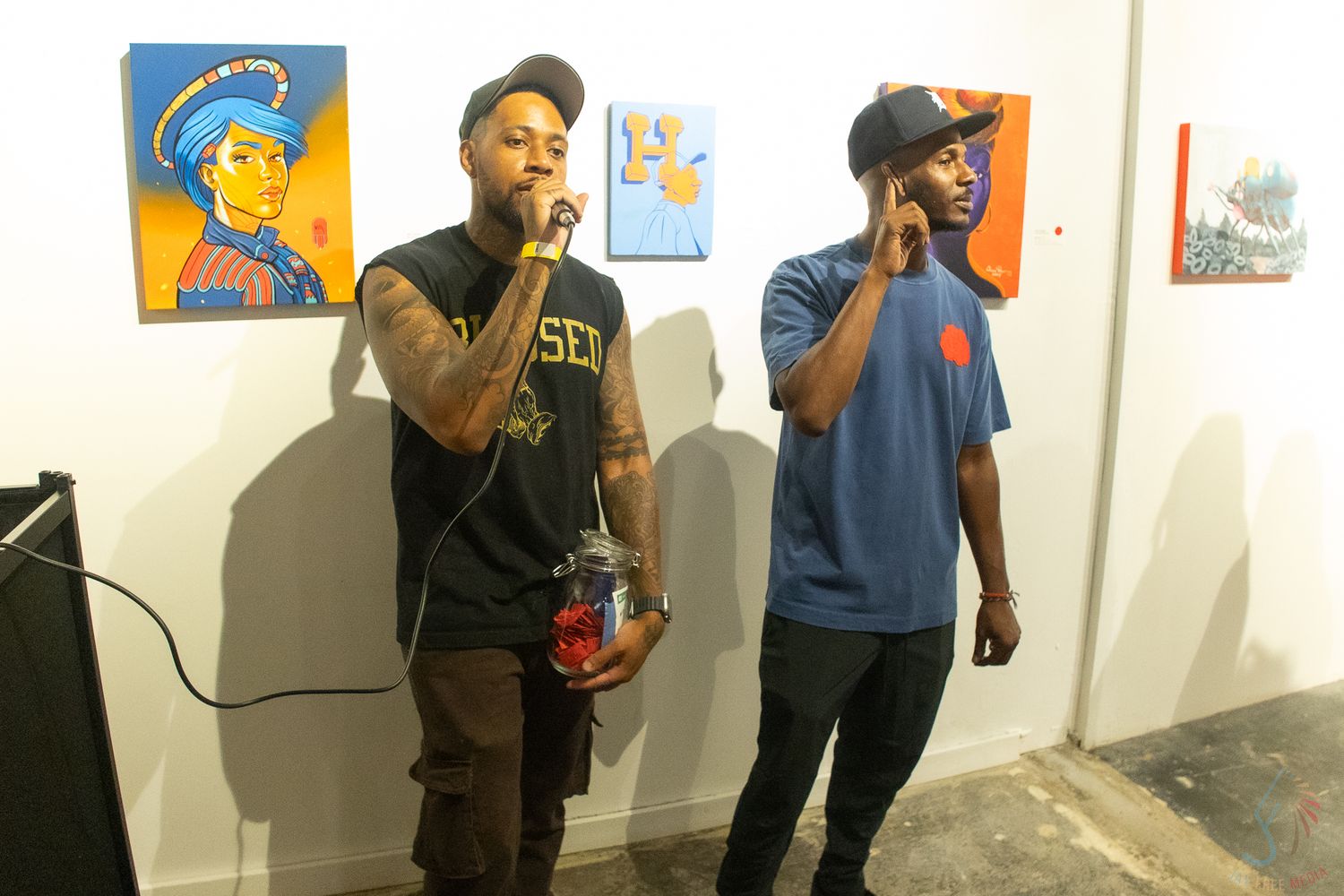 Two men speaking at microphone in art gallery with colorful paintings displayed on wall behind them.