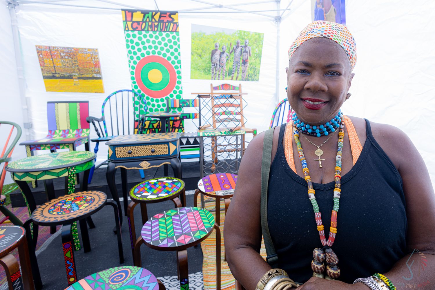 Black woman in black dress with colorful beaded jewelry stands beside painted stools at an outdoor art fair booth.