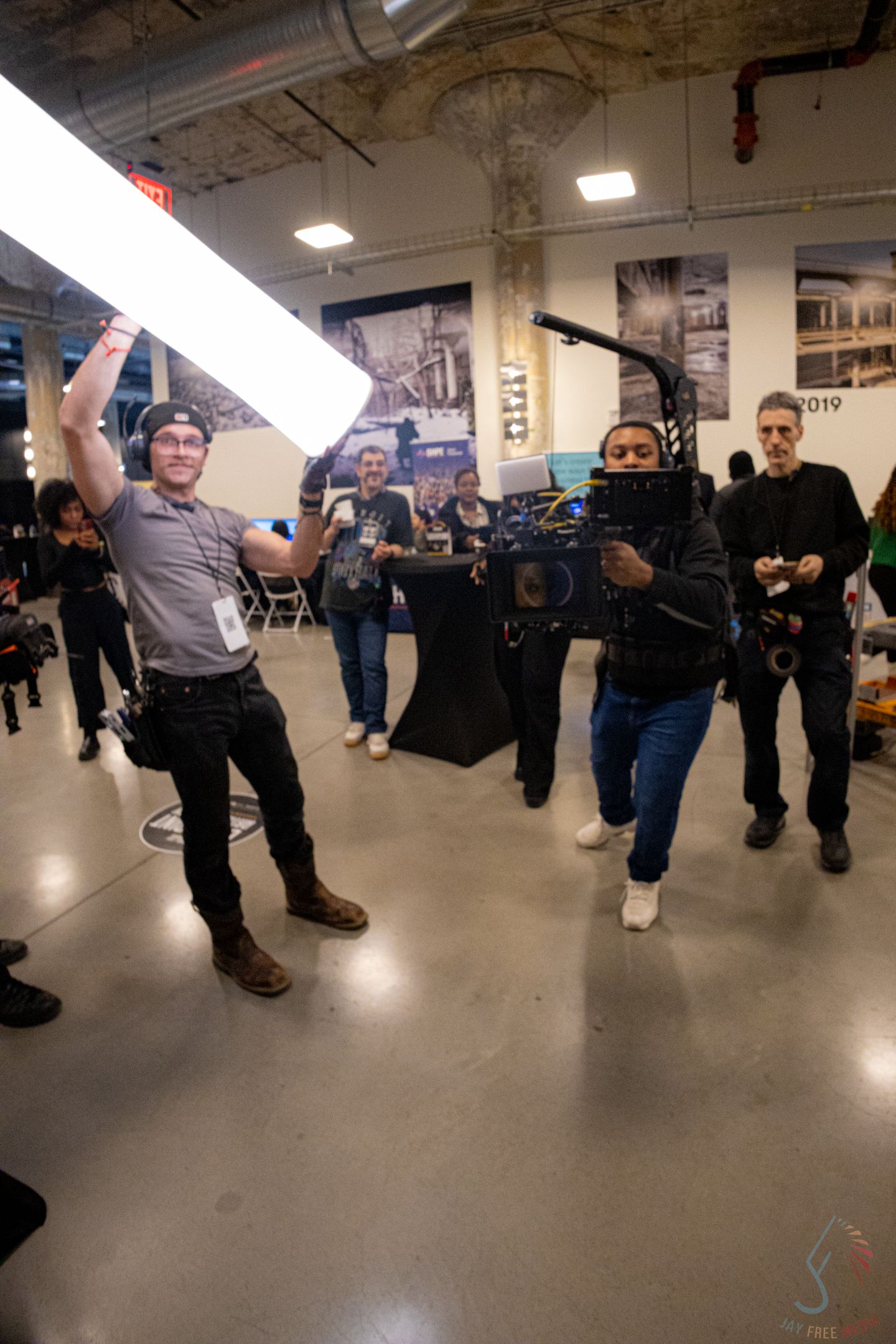 Two crew members hold film equipment including a camera and LED light panel at an indoor film industry event.